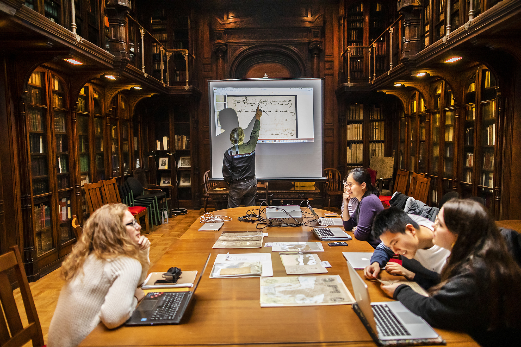 Professor pointing to handwritten words on document projected on screen in library with five students around long wooden table.