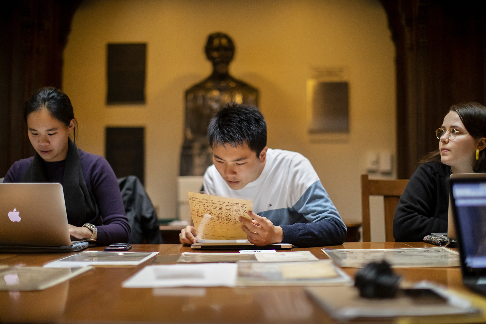 Student looking closely at a rare document in a plastic sleeve he is holding, while seated in a library.