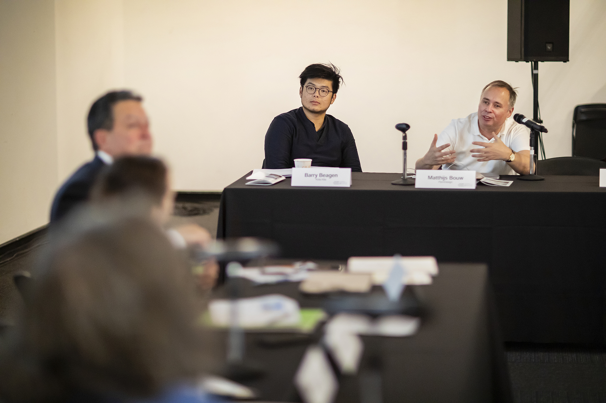 in a conference setting, seated presenters listen as one makes a point, gesturing for emphasis.