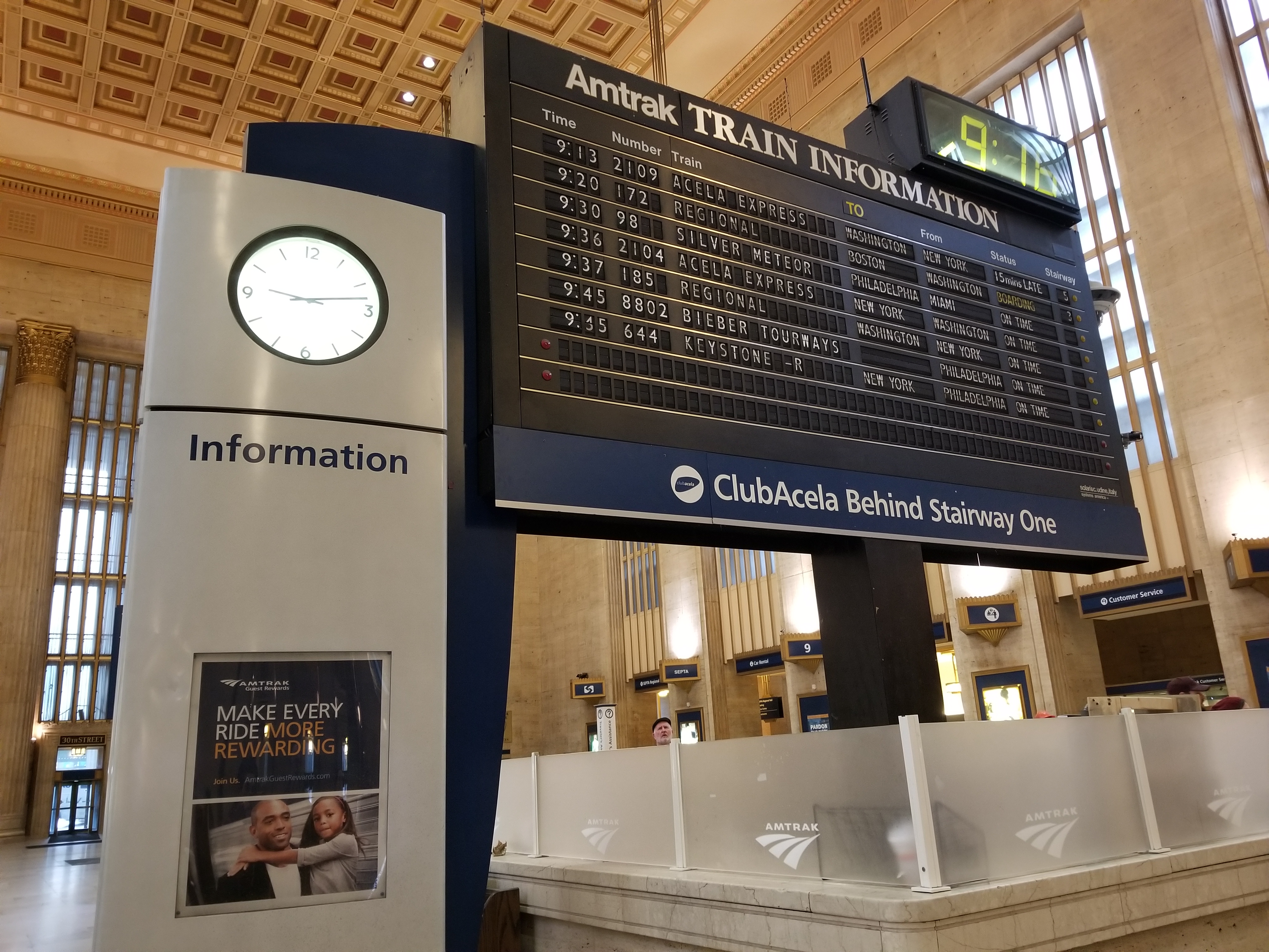 Split-flap board inside 30th Street Station surrounded by passengers and a clock