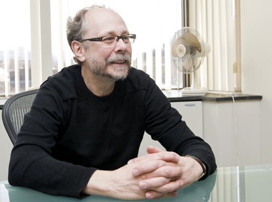 A man in a black shirt and wire-rim glasses sitting at a table with his hands crossed.