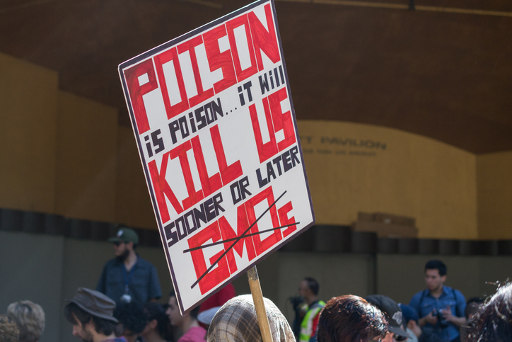 GMO protester holding a sign that reads "poison is poison...it will kill us sooner or later" and the letters GMOs crossed out