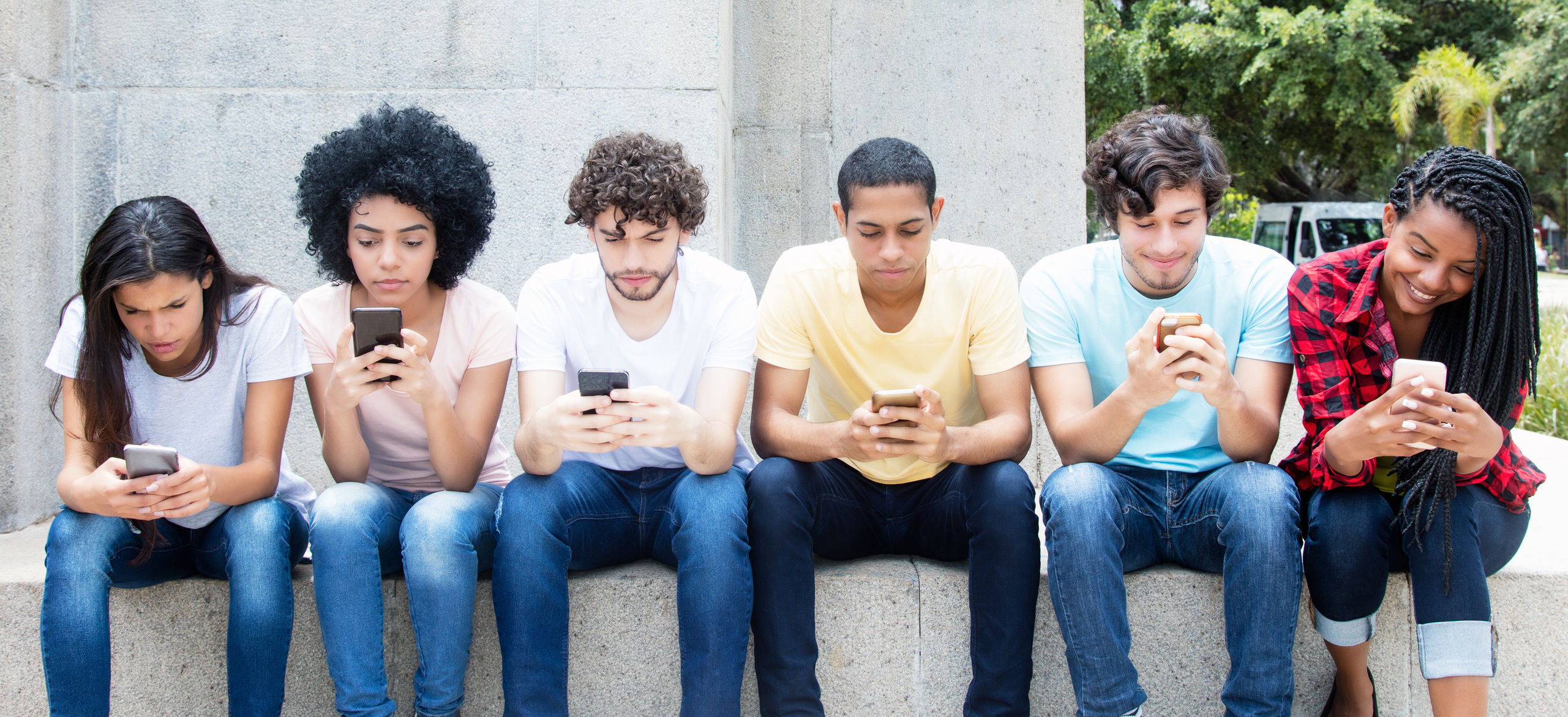 Teenagers of various races and genders sitting on a stone wall with cell phones in hand. 