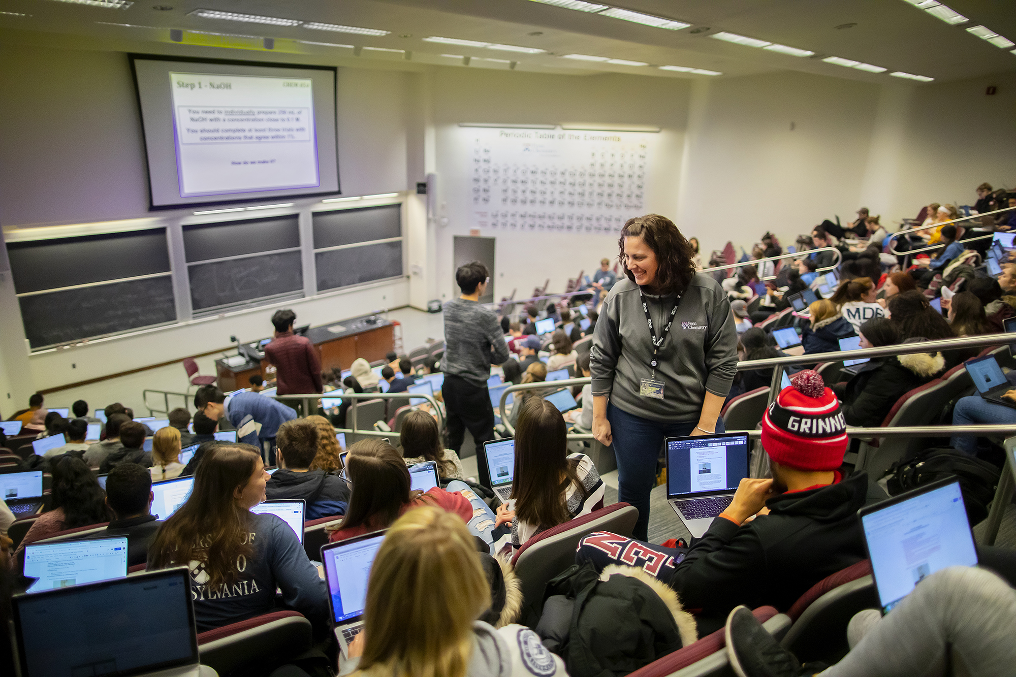 currano and two teaching assistants talk to students in a lecture hall while they work on their laptops