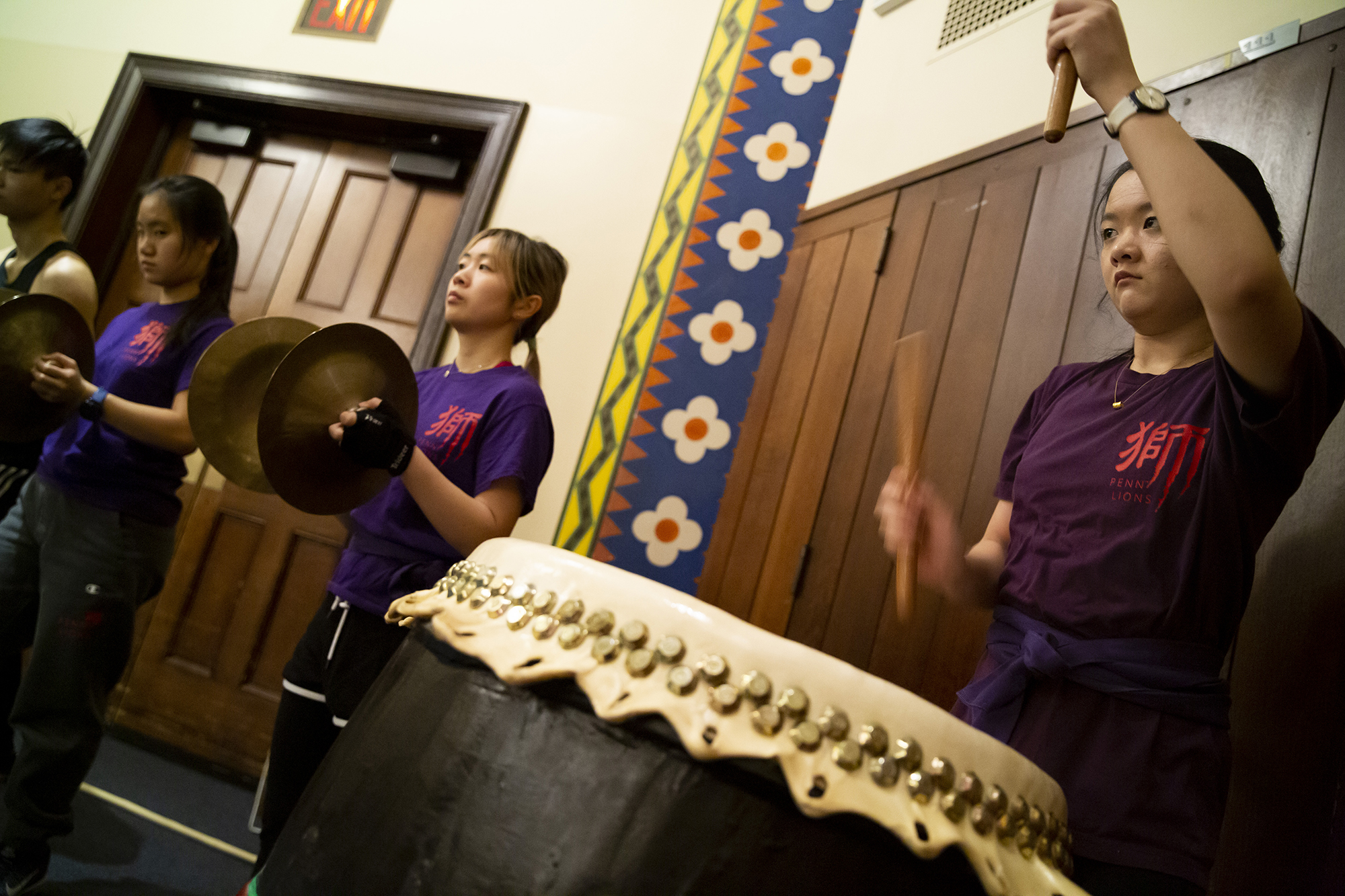 One student with arms raised while playing a drum and three other students on her left playing the symbols.