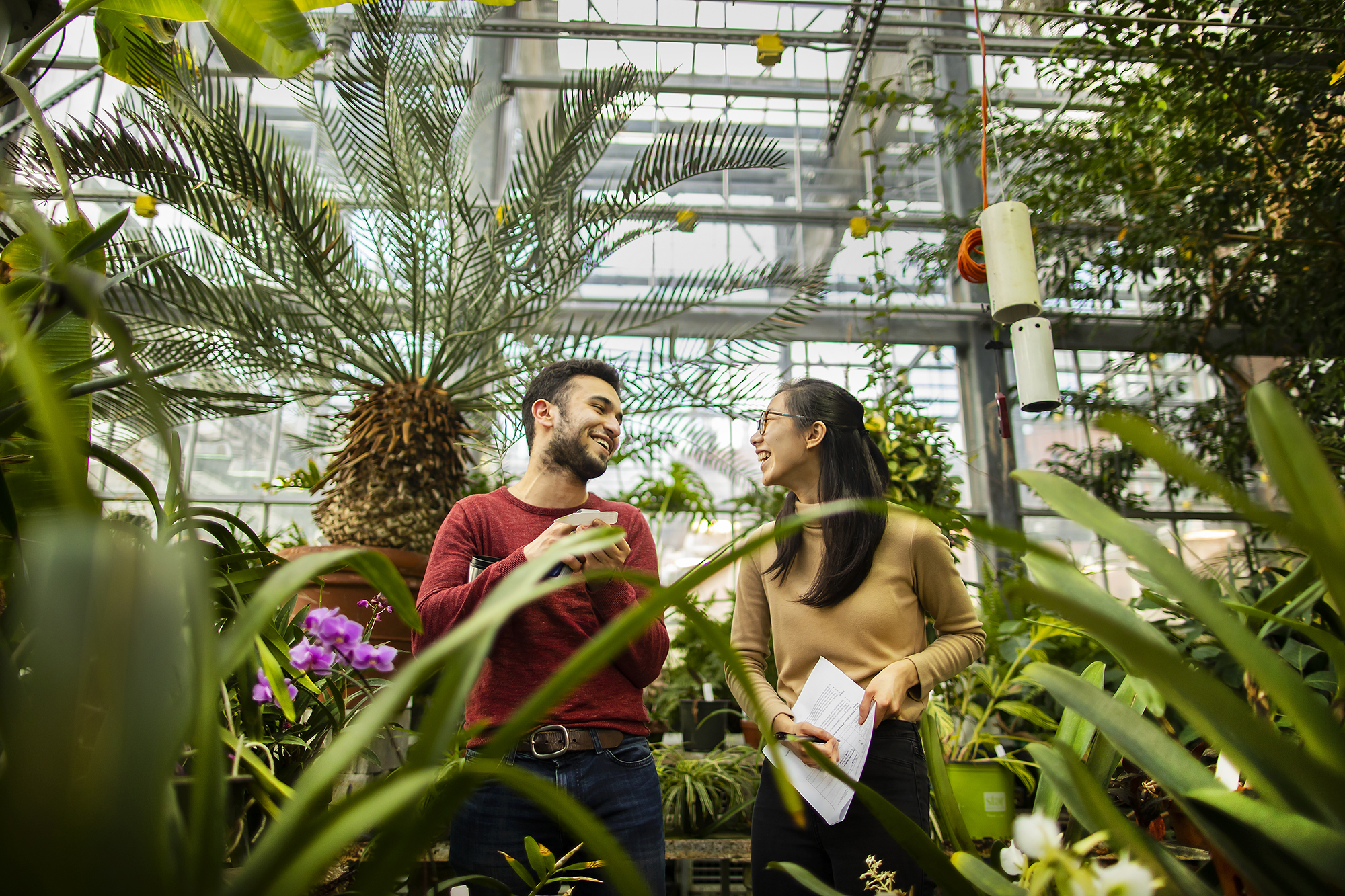 Two people smile at one another framed by plants in a greenhouse. One holds paper she is writing on.