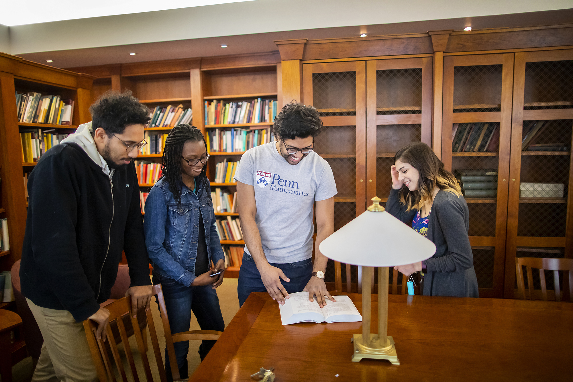 four students standing together in a library looking at a book on a table