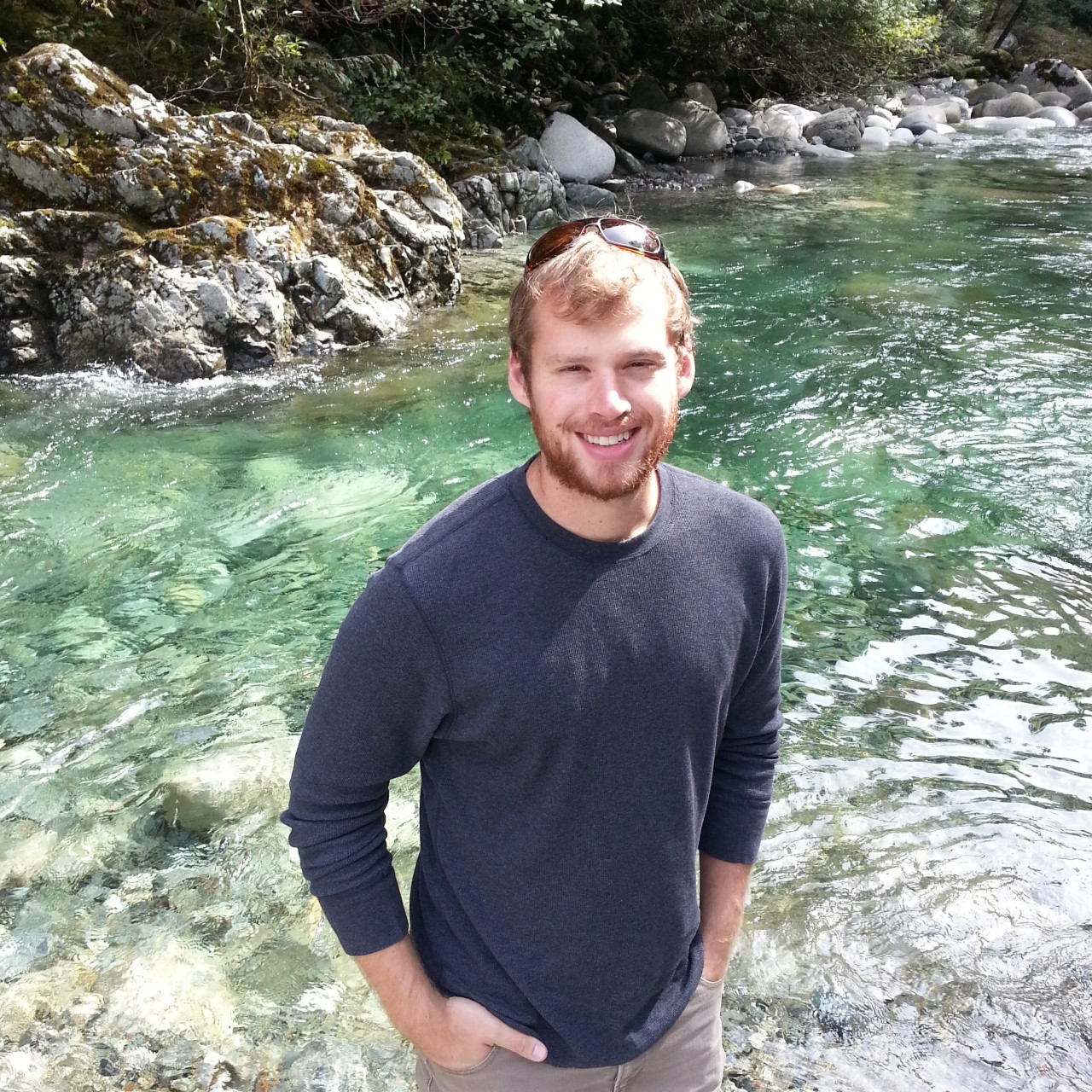 person smiling against a background of a clear water and rocky shores.