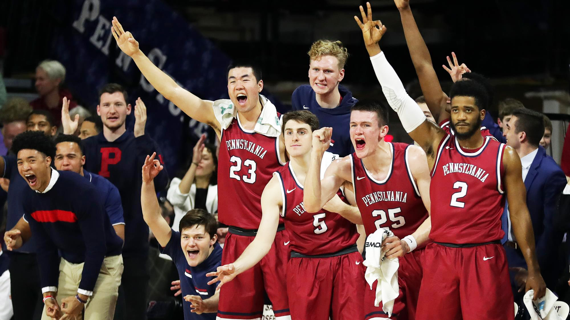 Men's basketball players celebrate on the bench holding up three fingers.