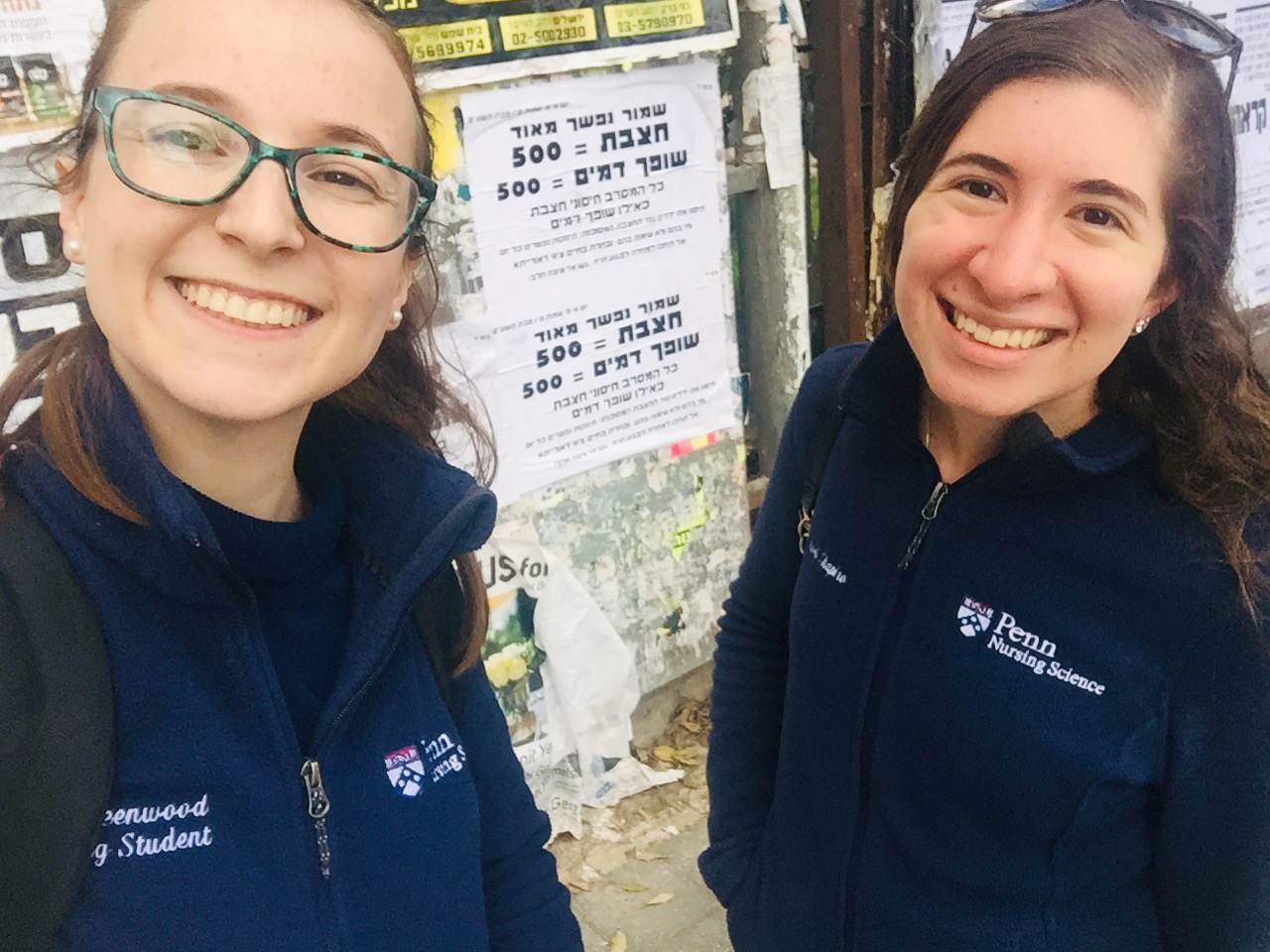 Two girls in blue Penn Nursing fleeces standing in front of a poster in Hebrew. 