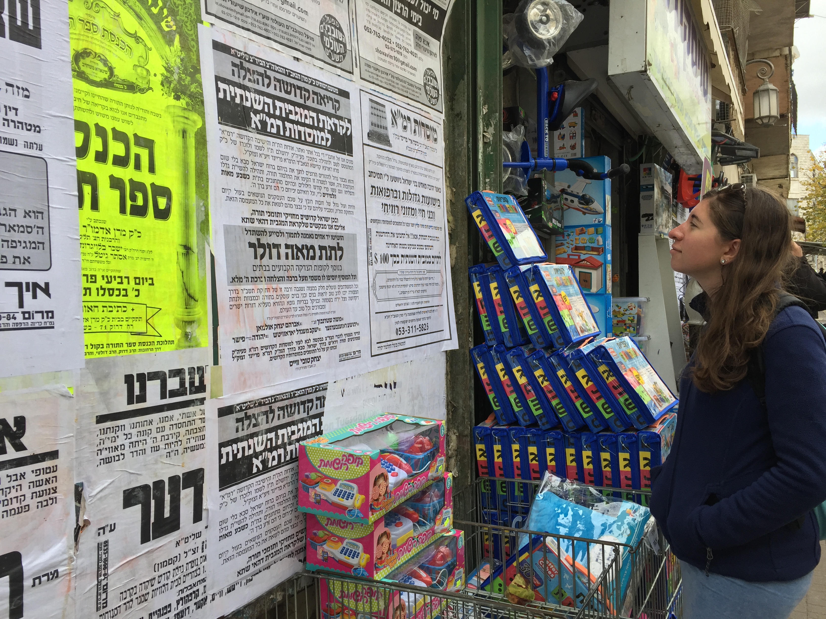 A girl in a blue fleece standing in front of a wall of black and white posters in Hebrew. Boxes of children's toys are in front of the wall.