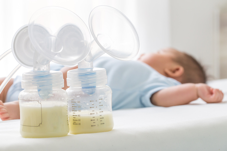 two bottles of breast milk with pump flange and infant in background