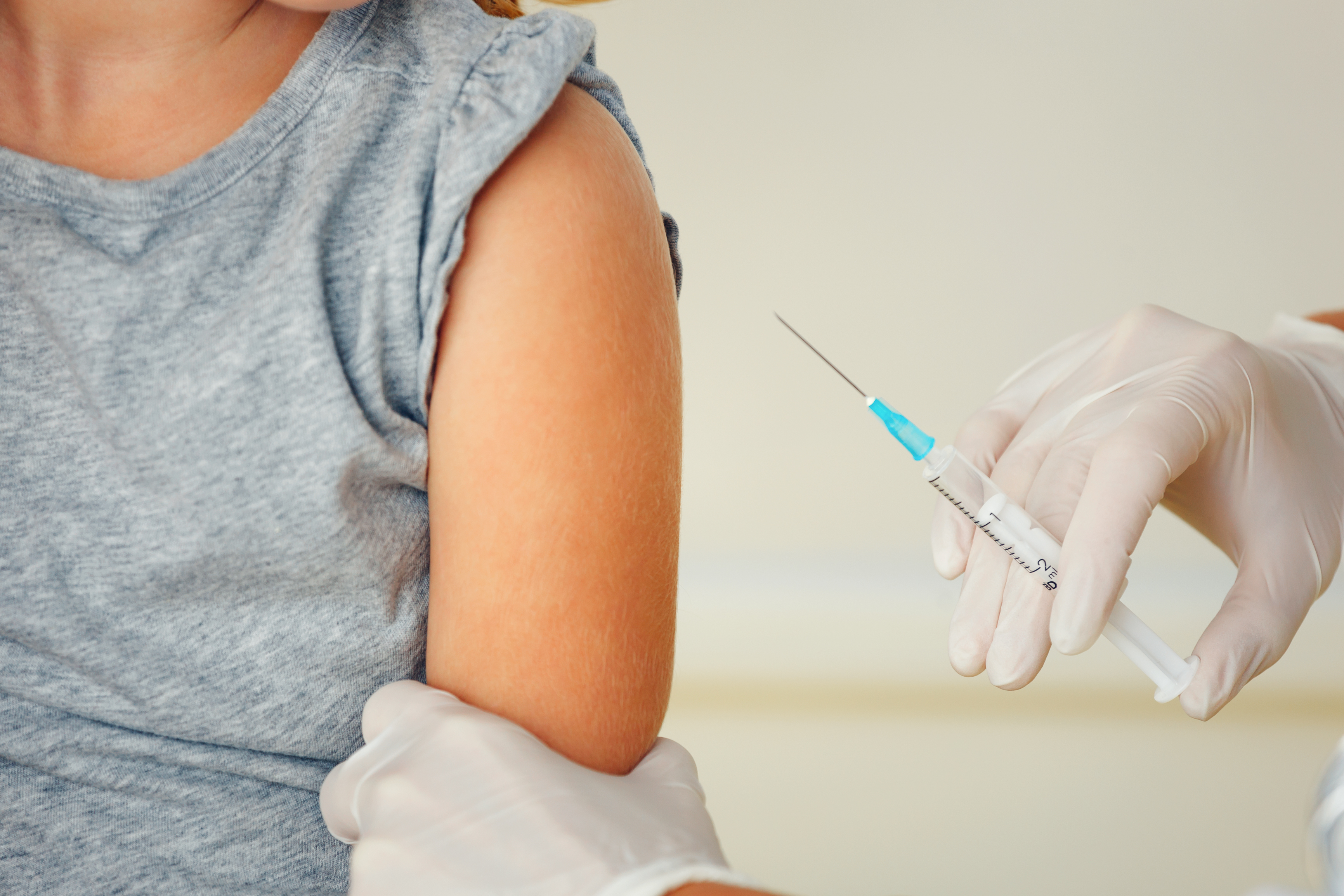 Child in a gray shirt sitting waiting to get a shot by gloved hands. 