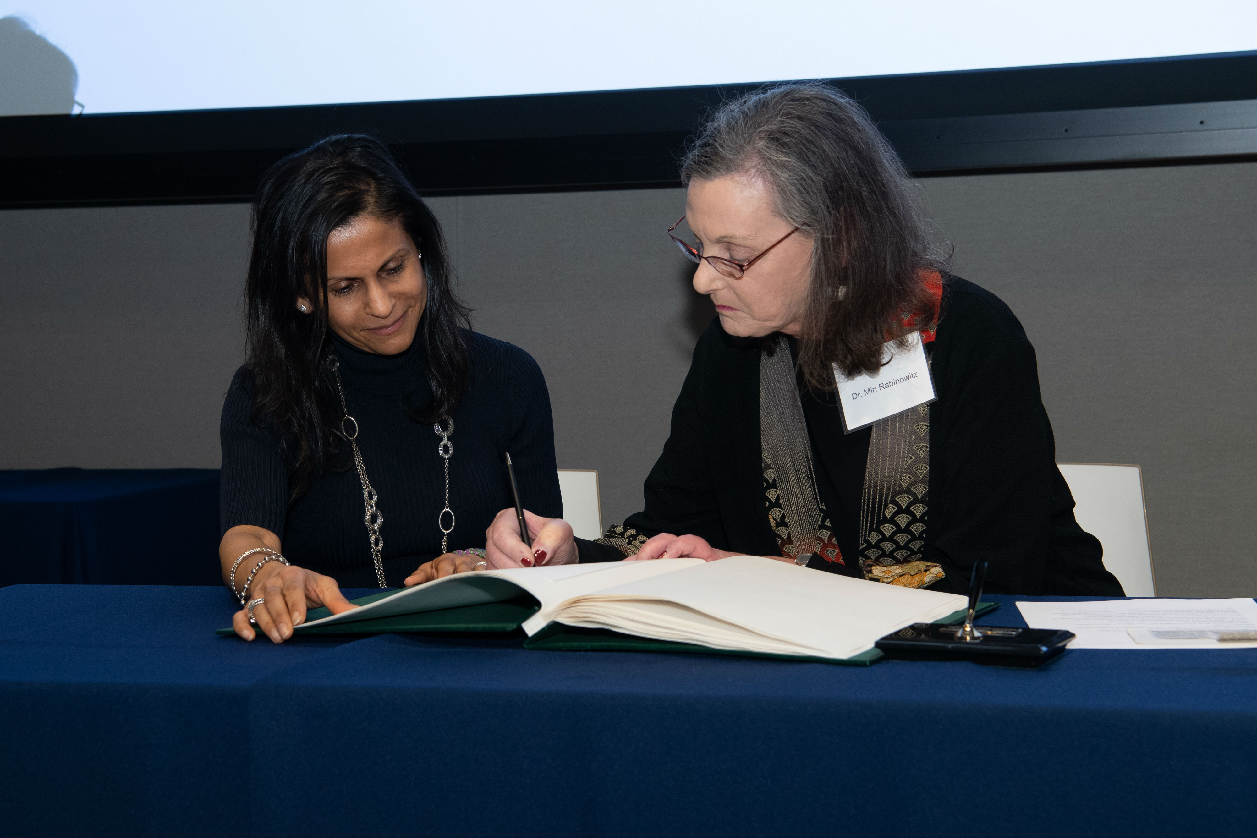 Two women sitting at a table, one signing a certificate. 