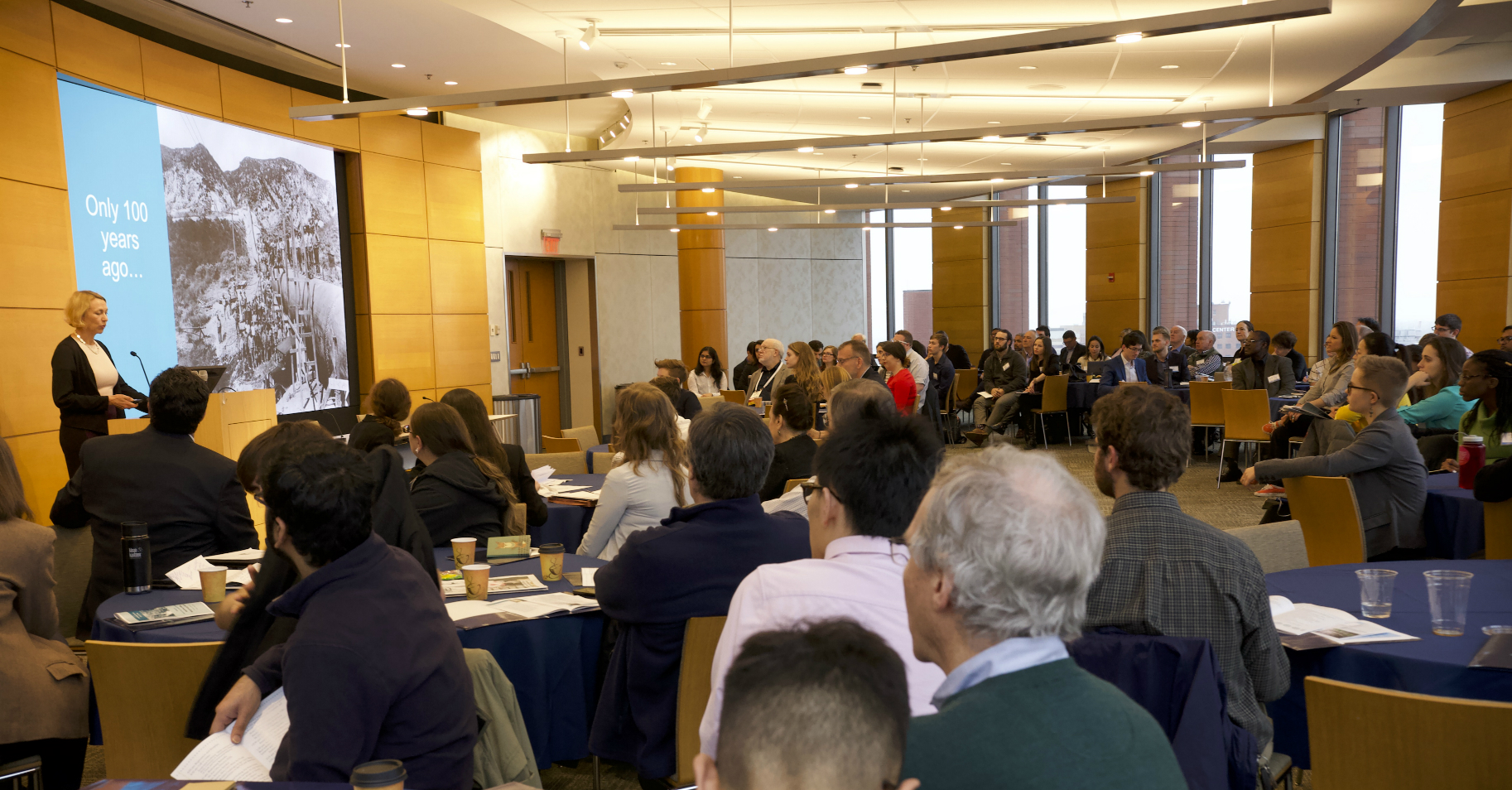 A speaker addresses a room full of people sitting at tables. A historic photo is displayed on a screen.