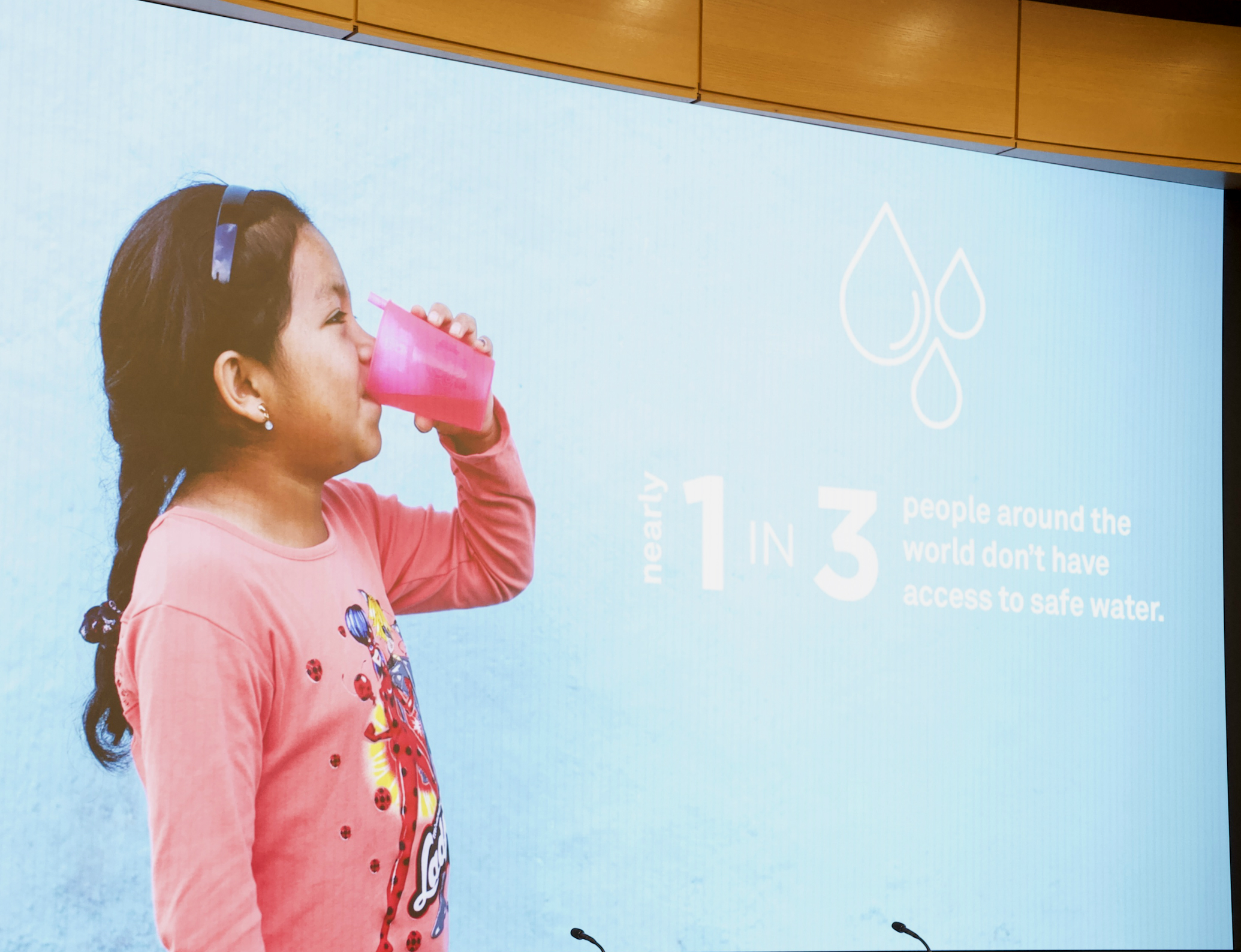 A projected image of a girl drinking water from a pink cup, alongside the words "nearly 1 in 3 people around the world don't have access to safe water"