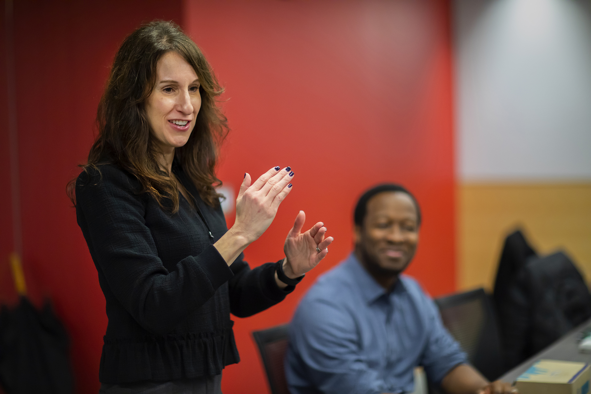 Woman in a black suit standing, gesturing with her hands, with an African American man in the background, in front of a red wall.