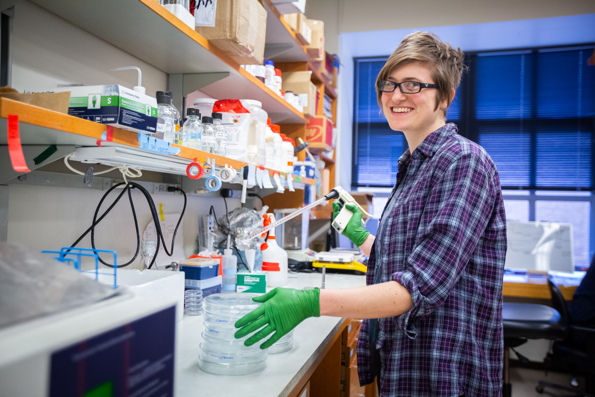 A person smiles while using a pipette and petri dishes in a scientific lab.