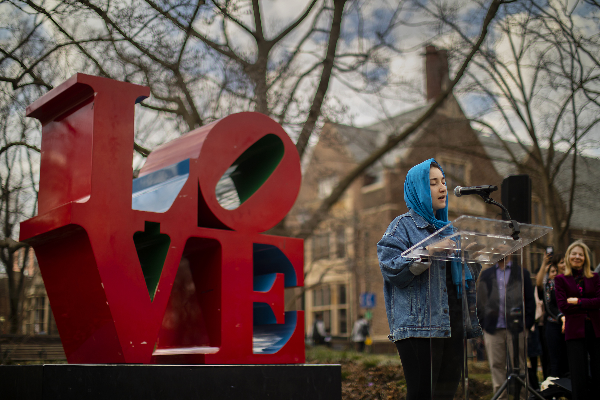 speaker at podium wearing head scarf addresses crowd while standing beside LOVE statue on campus