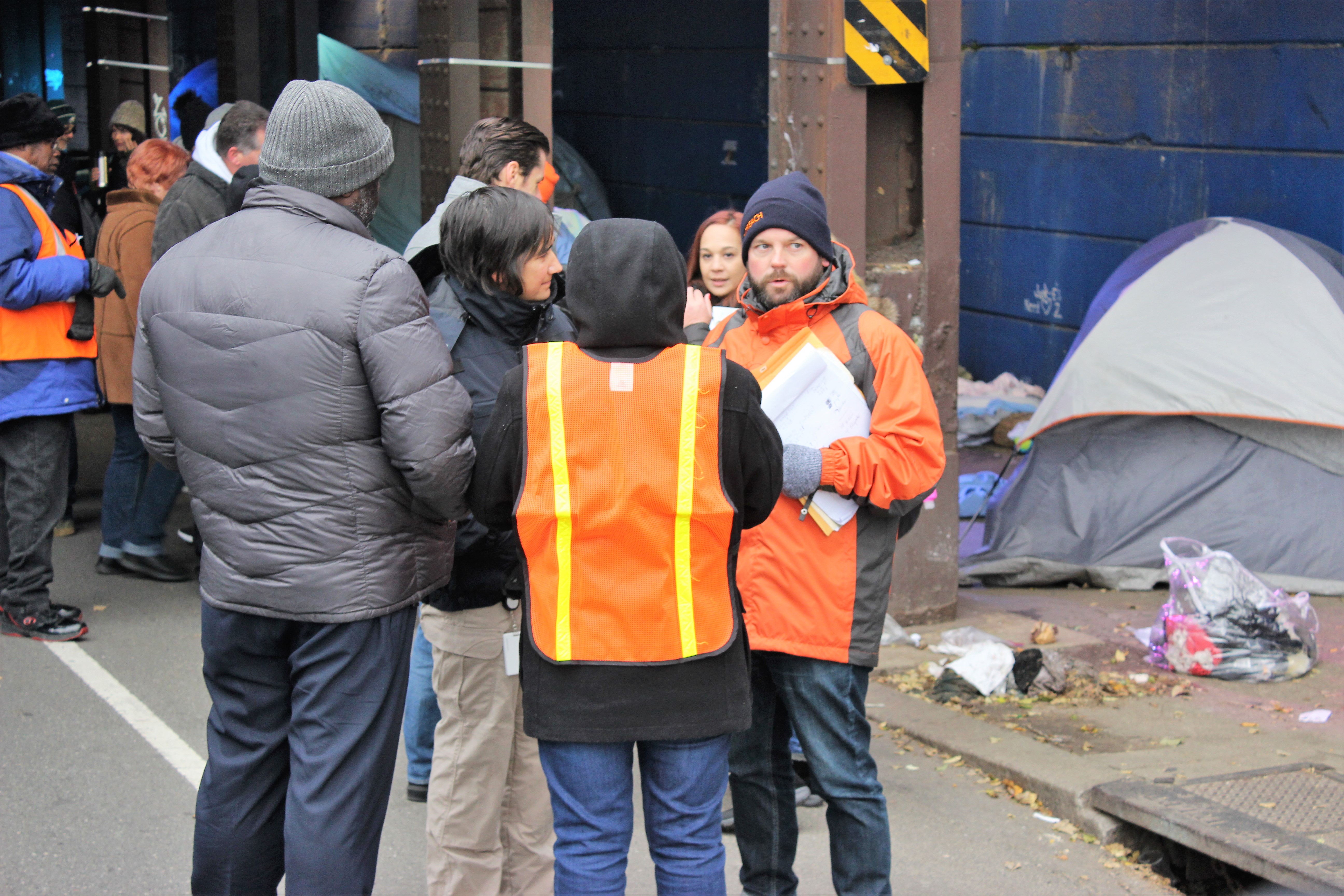 People standing in a group outside, with winter coats and orange vests, in front of a tent and plastic bags.