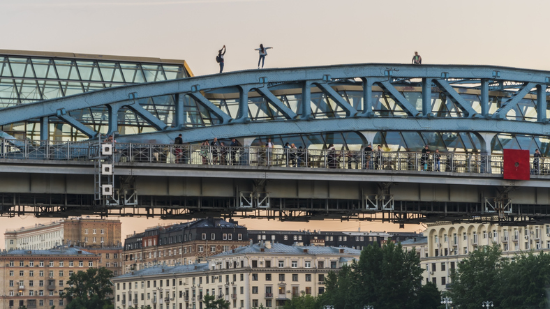 Two adolescents walking on the top structure of a bridge, a third sits, while people are on the walkway beneath the top of the bridge. 