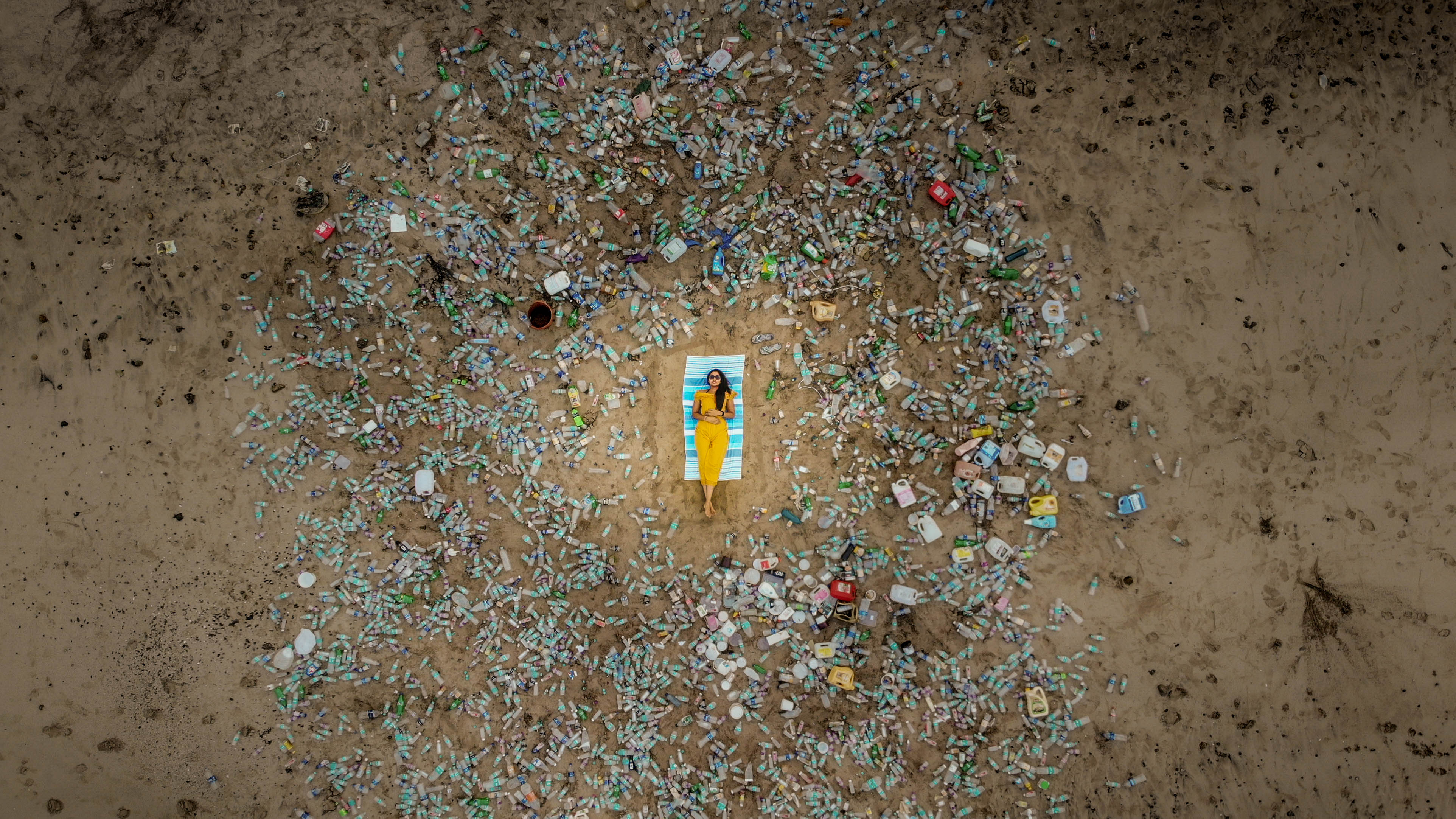 A person reclines on a chair on a beach surrounded by plastic bottles