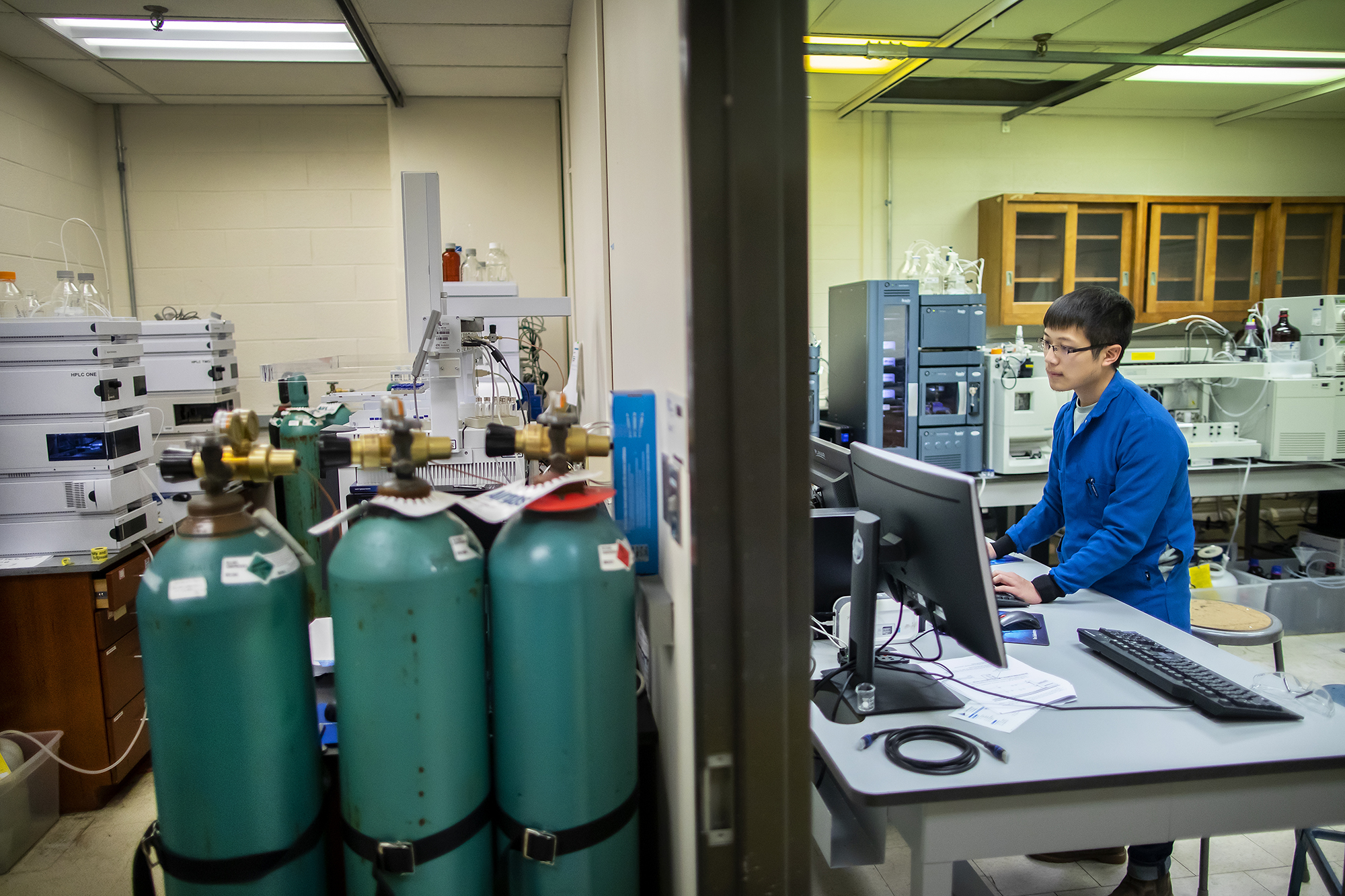 a man working on a computer inside a chemistry lab with equipment in the background