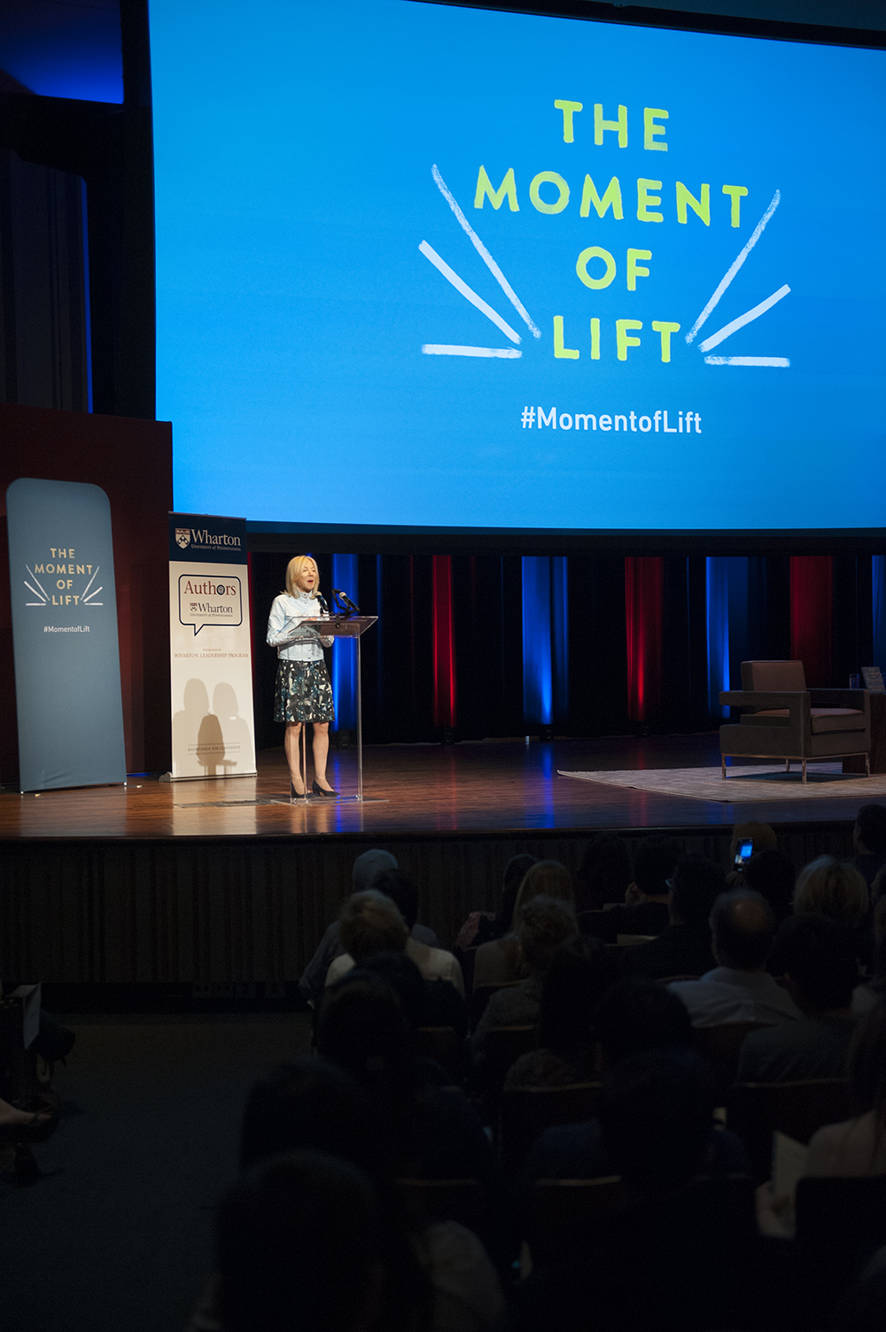 Amy Gutmann speaking on stage at a podium under a large screen reading #MomentofLift