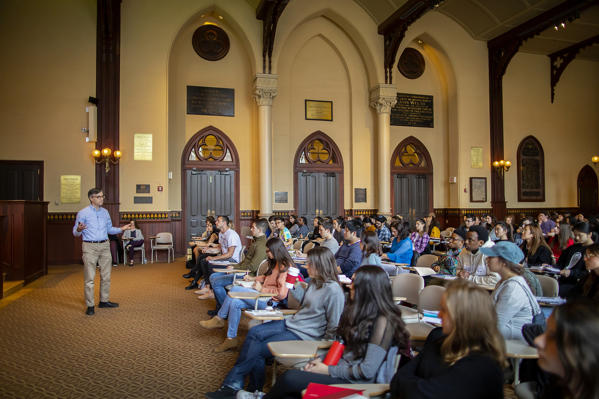 A man in a blue shirt and khakis standing in front of rows of students sitting at desks.