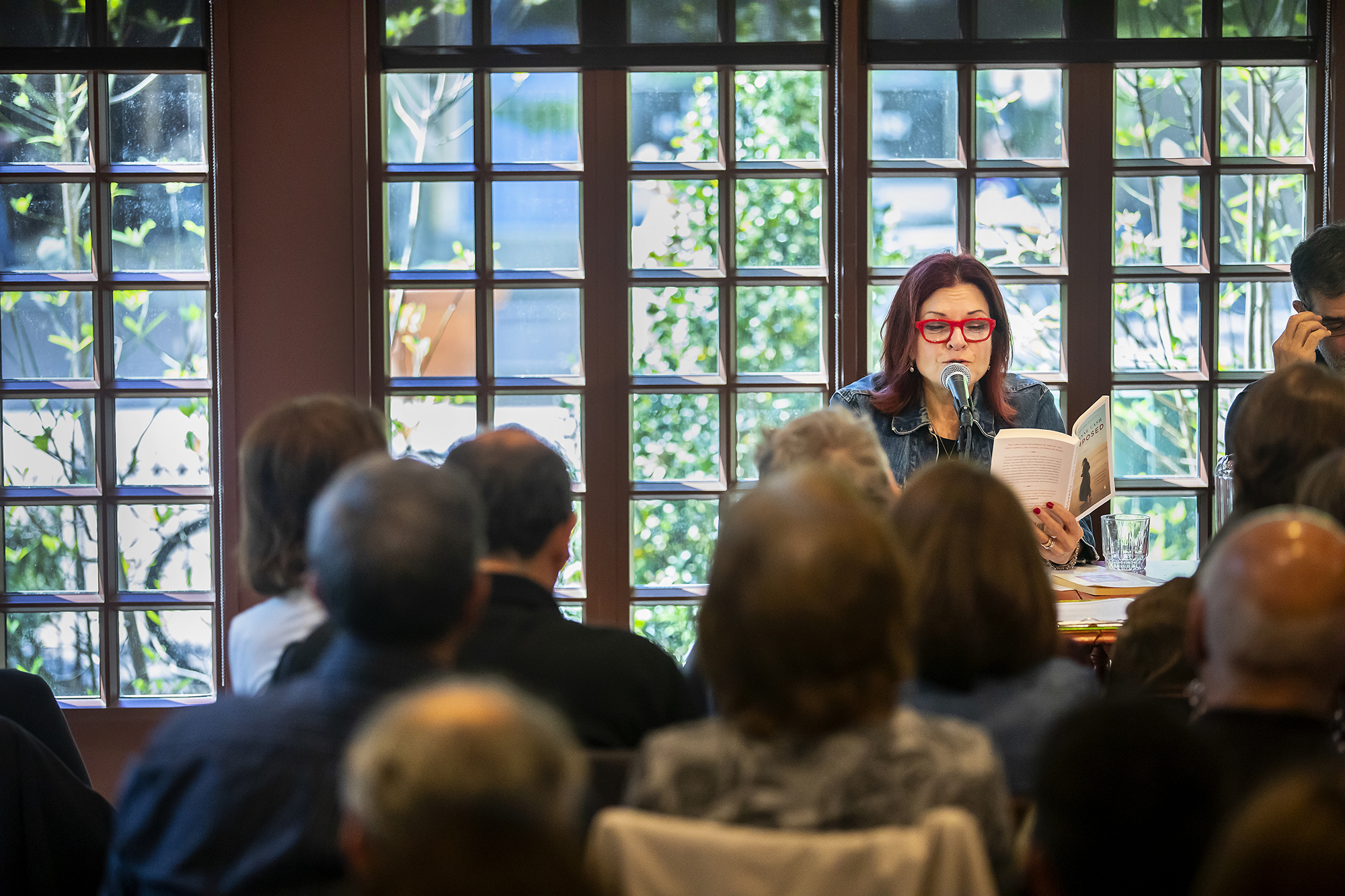 Rosanne Cash wearing red glasses reading her book while seated at a table in front of a crowd. 