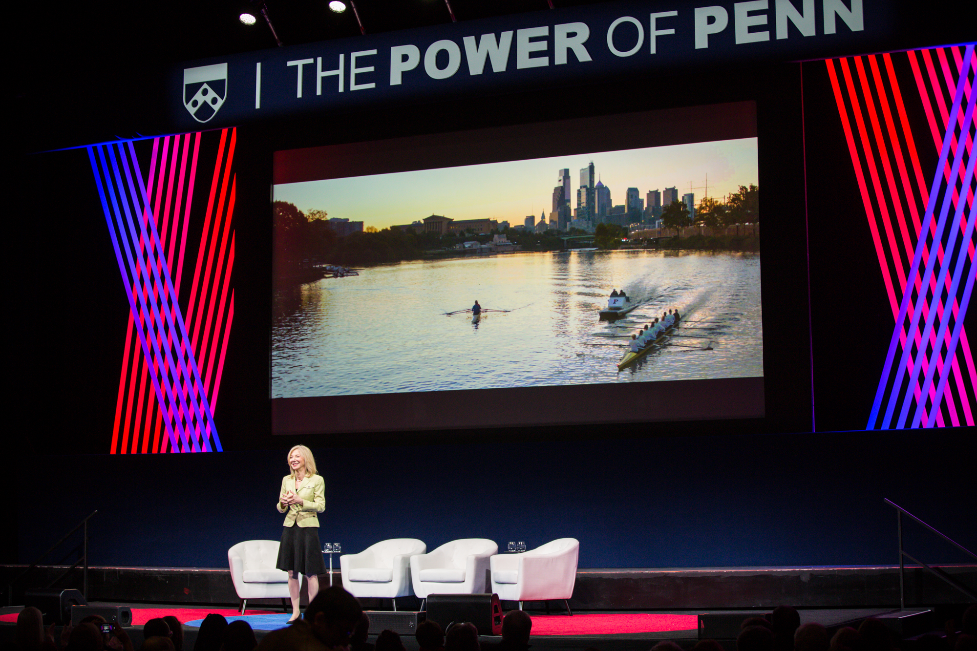 Amy Gutmann standing on stage with large screen behind her showing a video