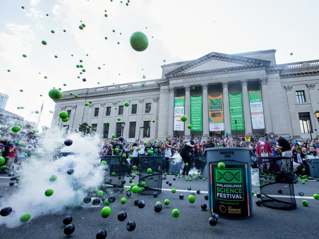 a large group of people in front of the Franklin Institute building with a science demonstration (with smoke and the aftermath of an explosion that caused colored balls to fly into the air) in the foreground