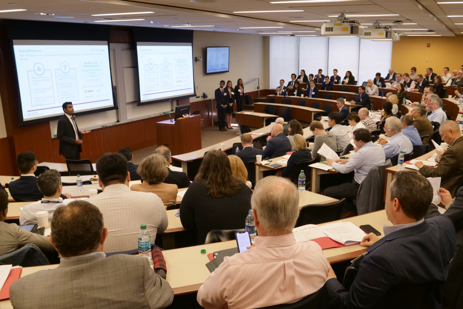 a person standing at the front of a full lecture hall giving a presentation