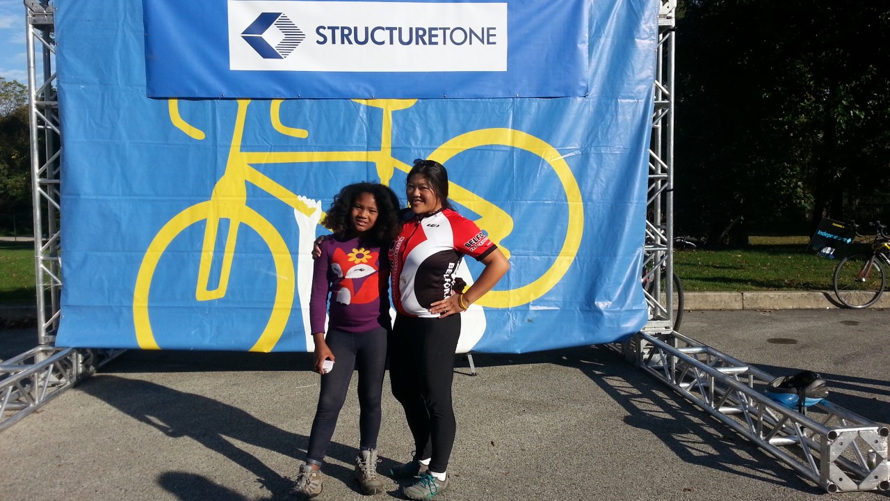 A child and adult wearing bicycling clothing pose in front of a banner displaying a bicycle