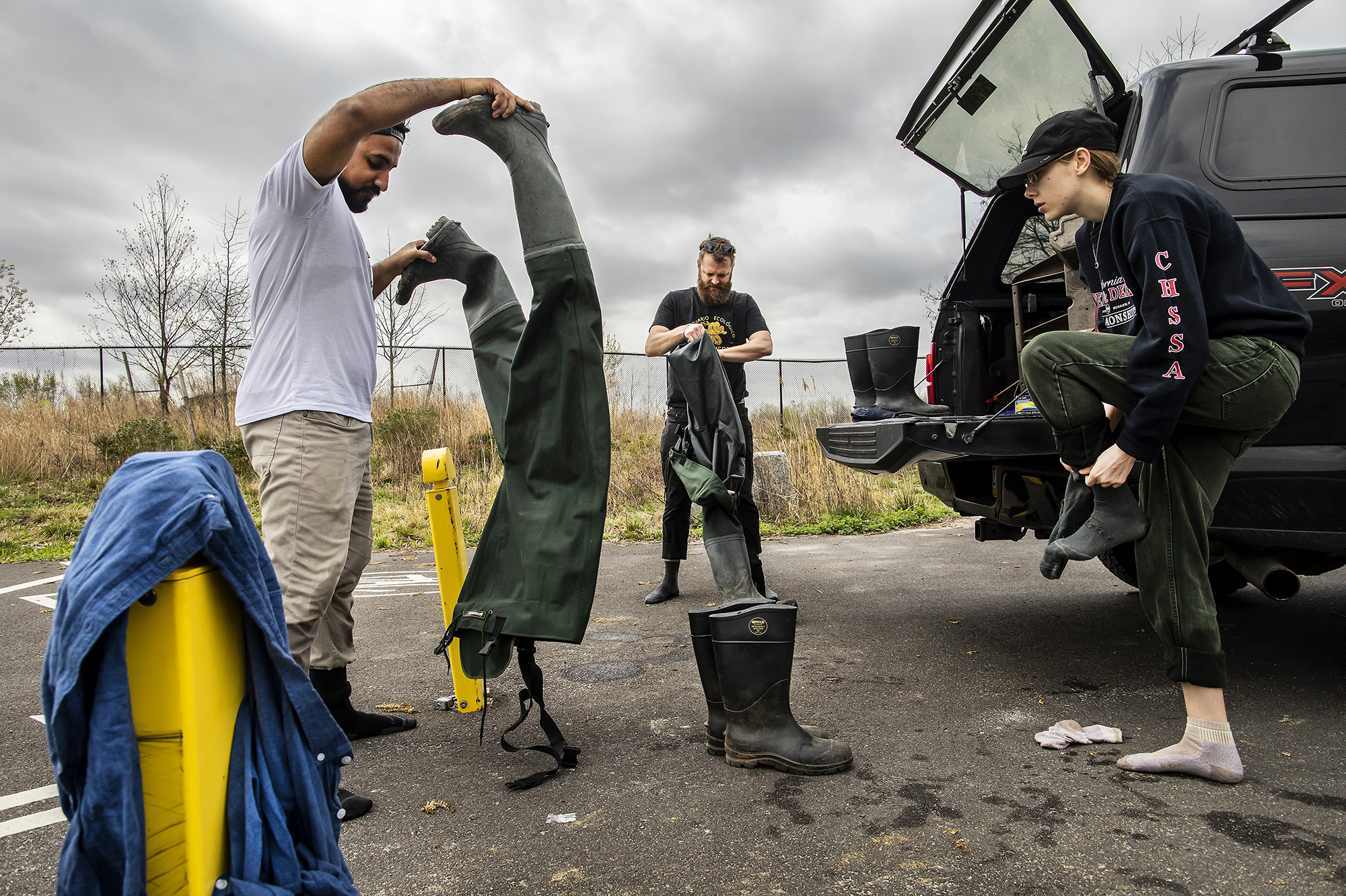 Three people take off hip waders by a black truck with the trunk open, one changing out of wet socks