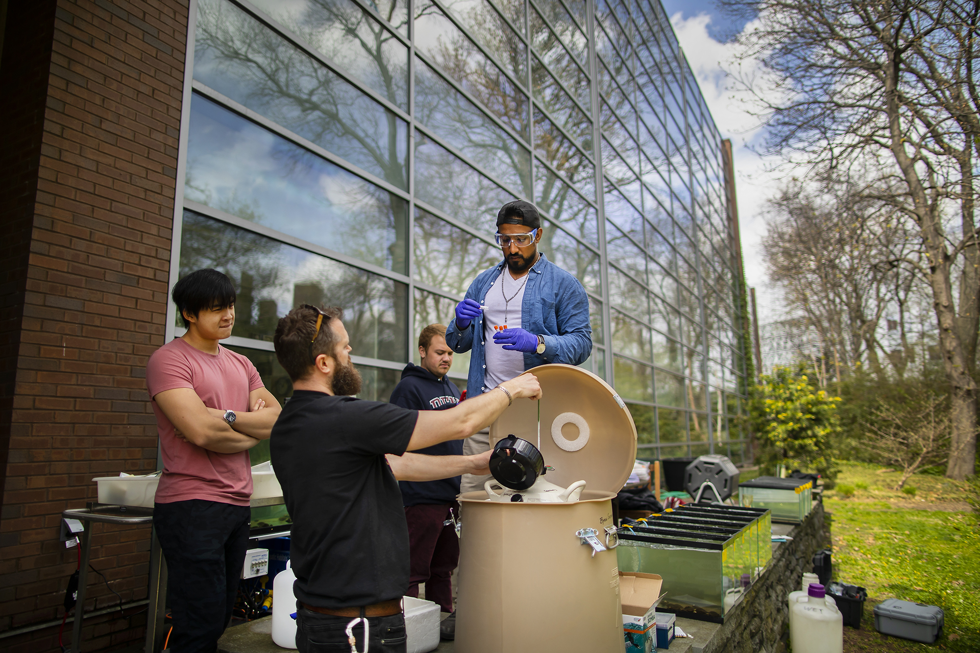 An instructor opens a large tank as students look on, one ready to place a sample inside. They are outside and the reflection of trees appears on a nearby building