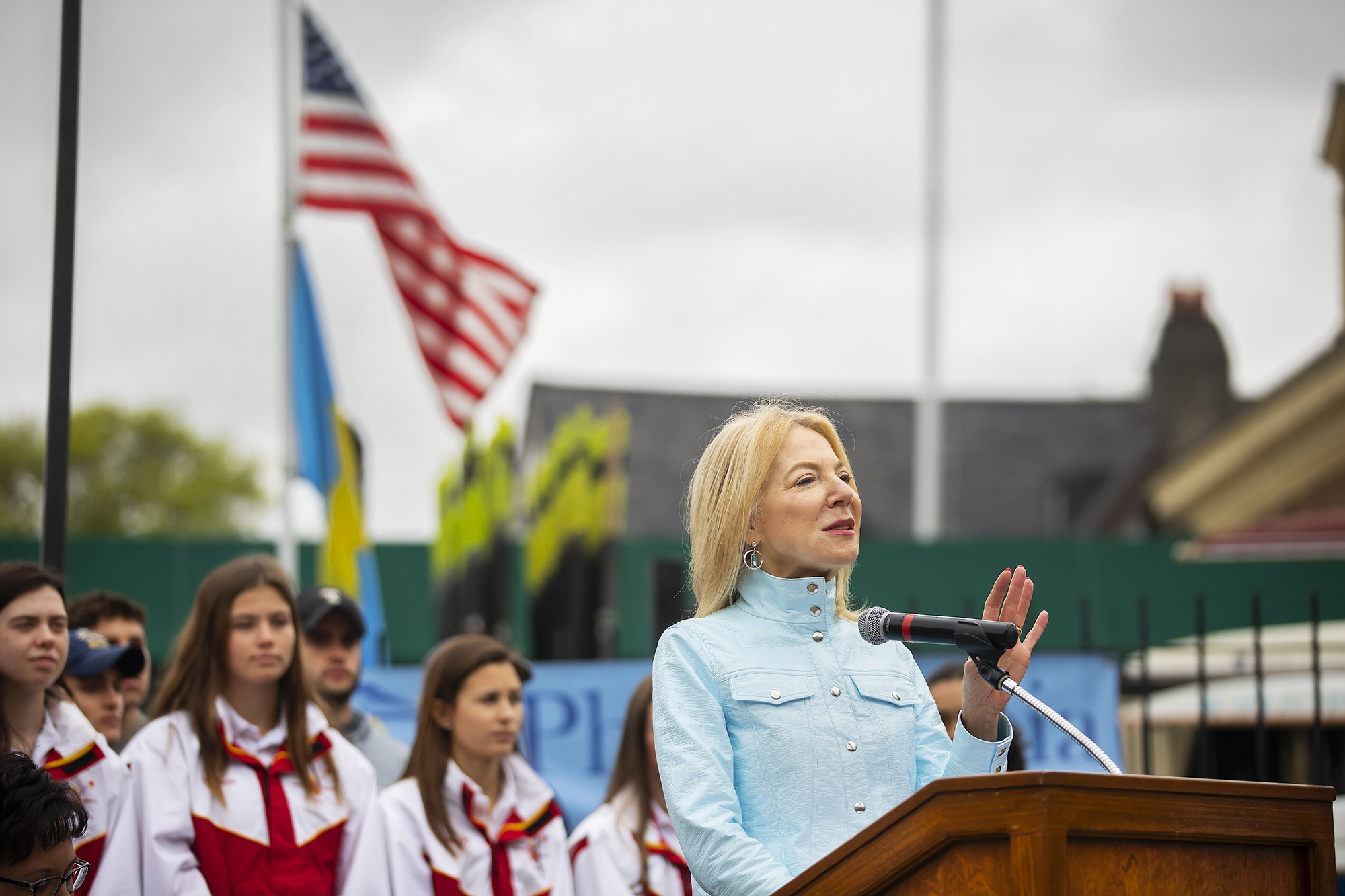Amy Gutmann speaks at a podium with American flag and a crowd behind her. 