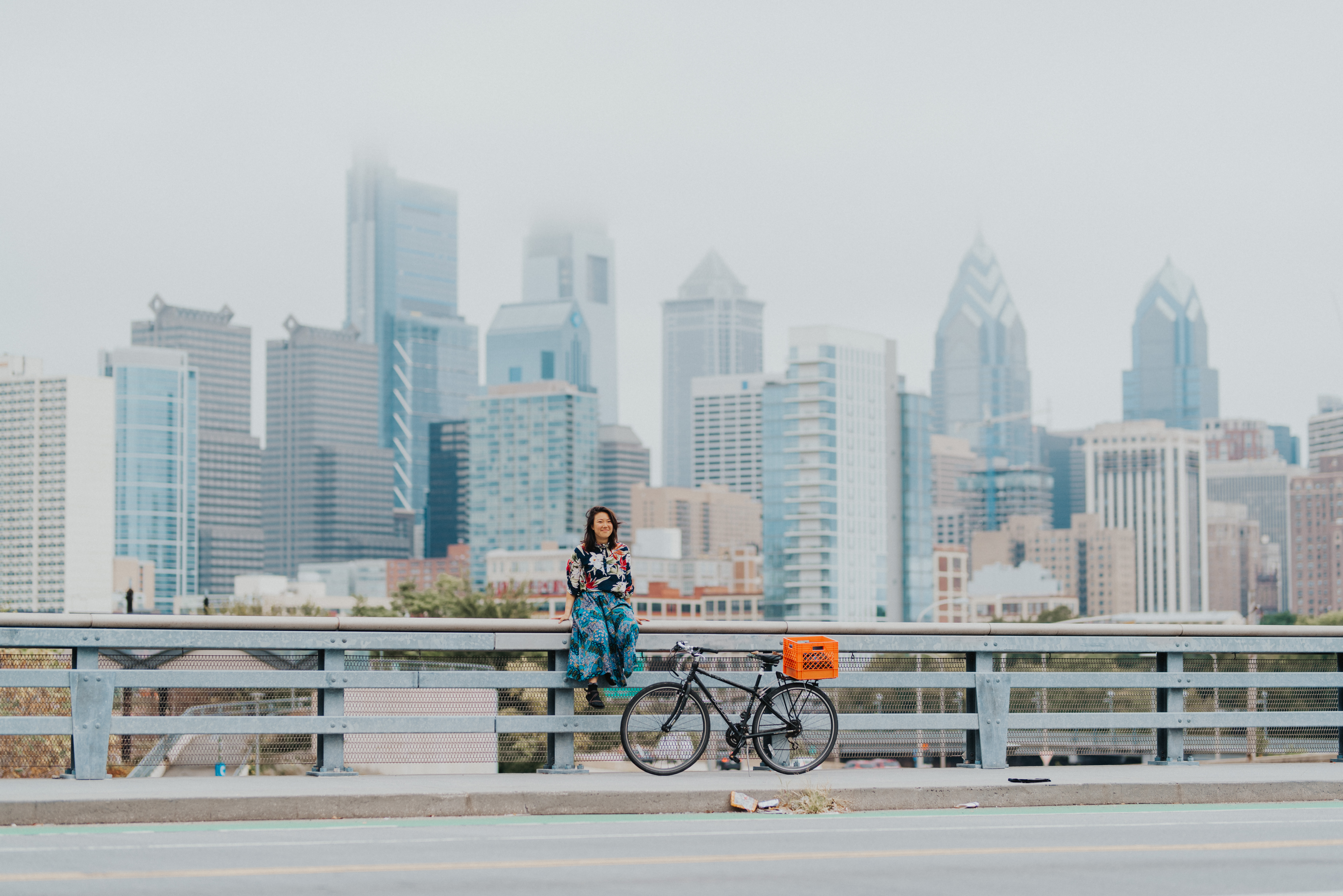 Student sitting on railing next to her bicycle, the Philadelphia skyline in the background.