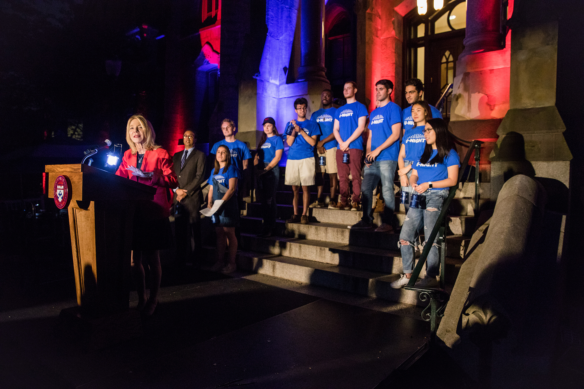 Amy Gutmann speaking at the podium with students on the steps of College Hall behind her.