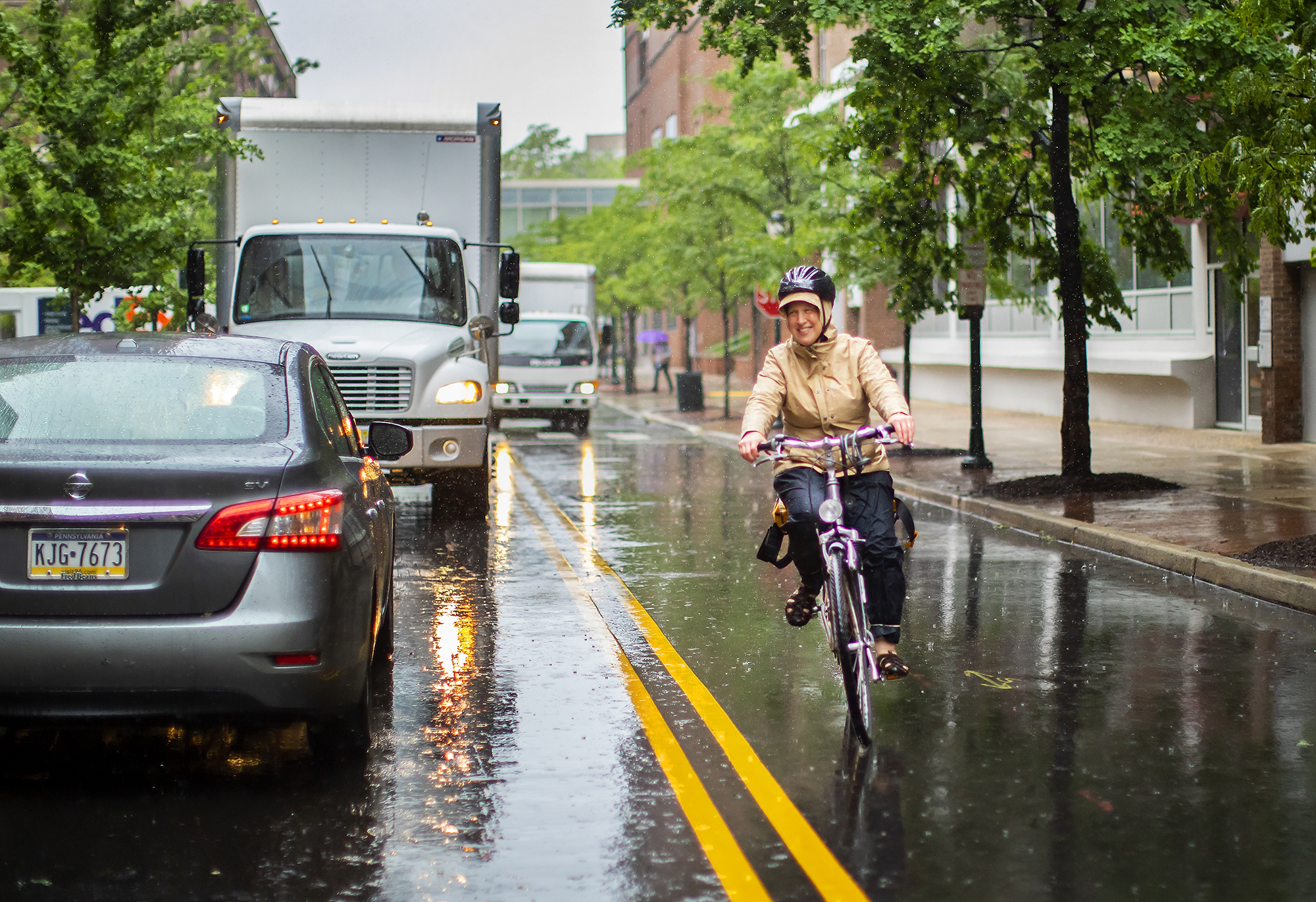 Bike rider in the rain coming down a street with cars and trucks in the opposite lane