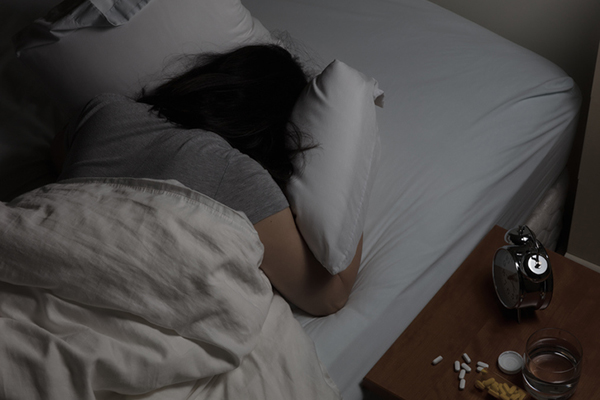 Person in bed with face in pillow and an alarm clock and pills on the bedside table.
