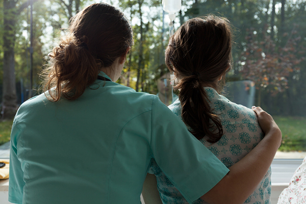 A hospital worker with arm around a patient seen from behind as they look out the window.