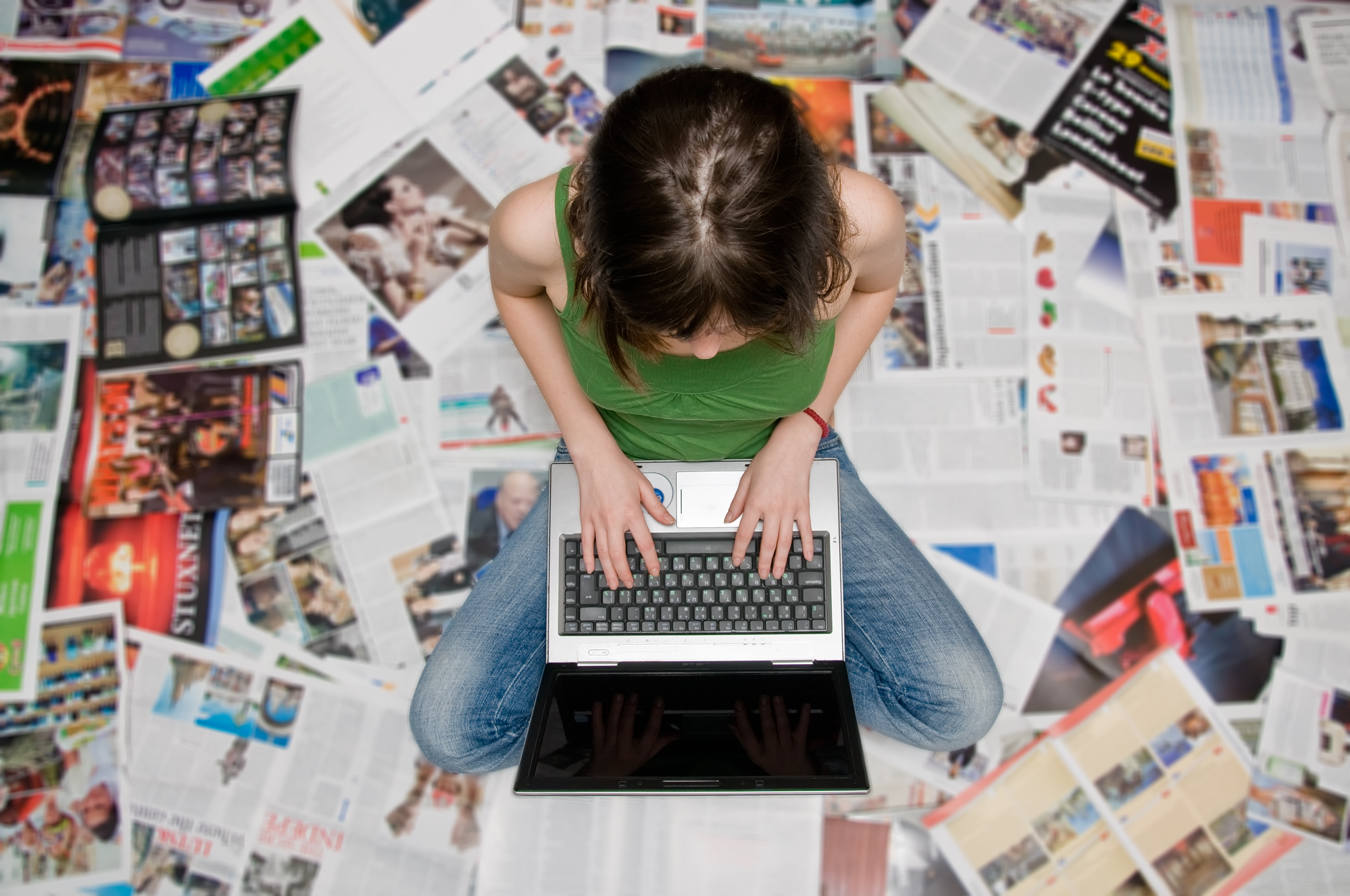 a person sitting on a stack of open magazines and newspapers working on a laptop