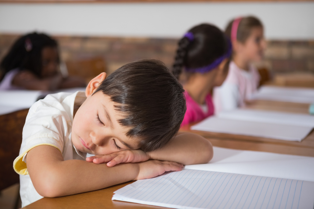 Child in a white shirt sleep on a paper-covered desk in a classroom, with three students in the background.