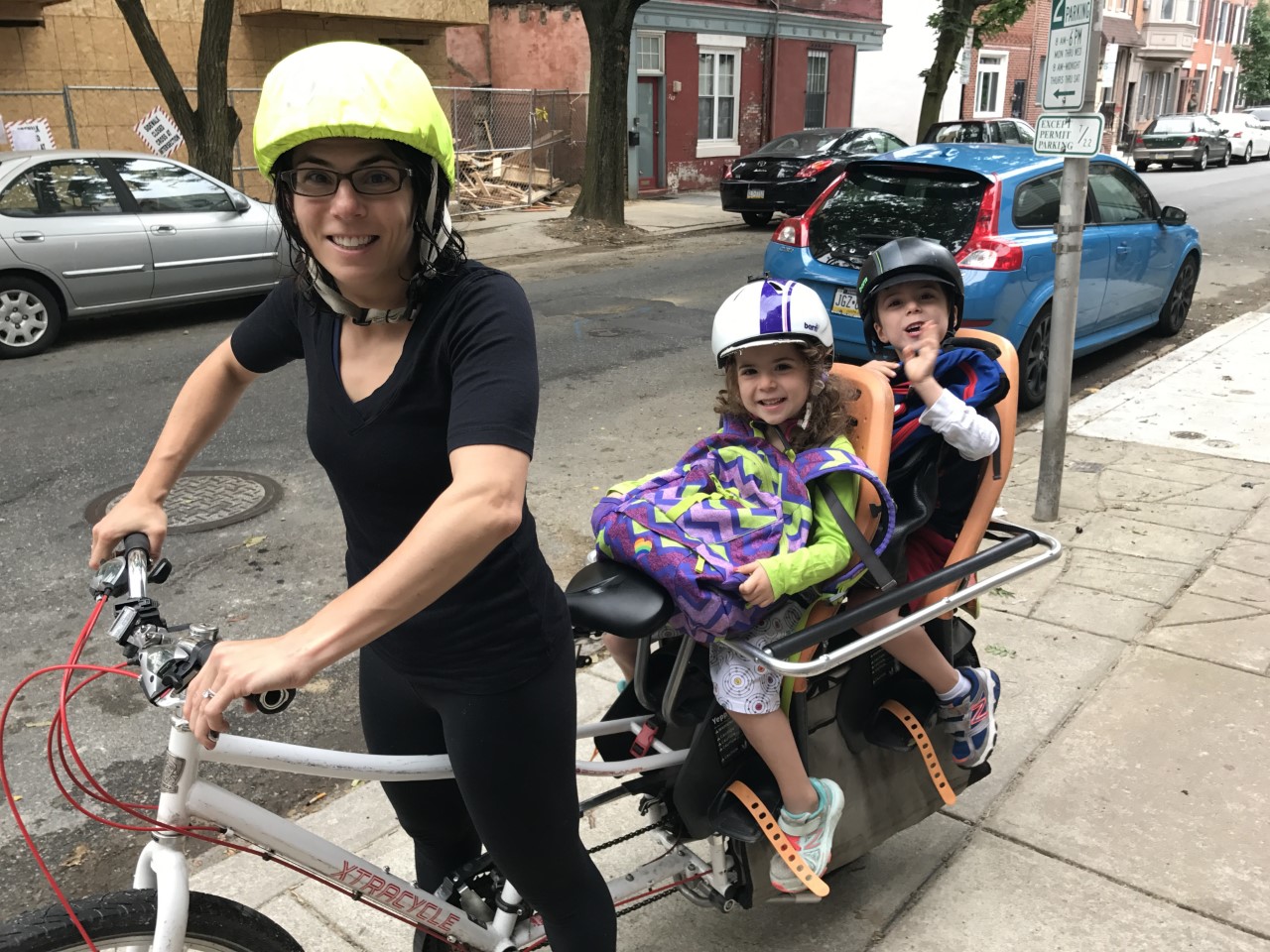 An adult with two children on a cargo bike post on the sidewalk
