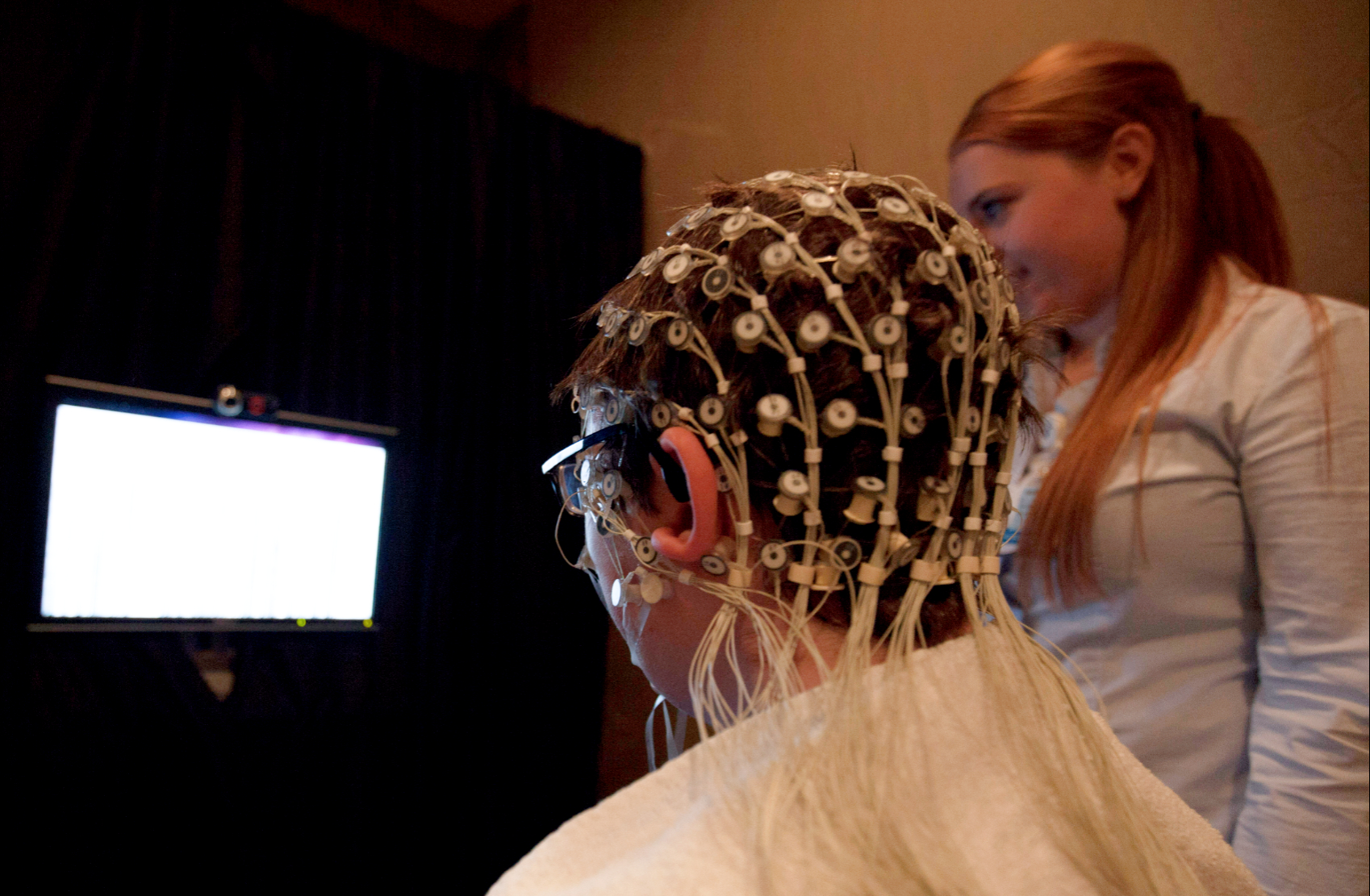 A child wearing an electroencephalogram cap looking at a bright screen, with someone standing nearby.