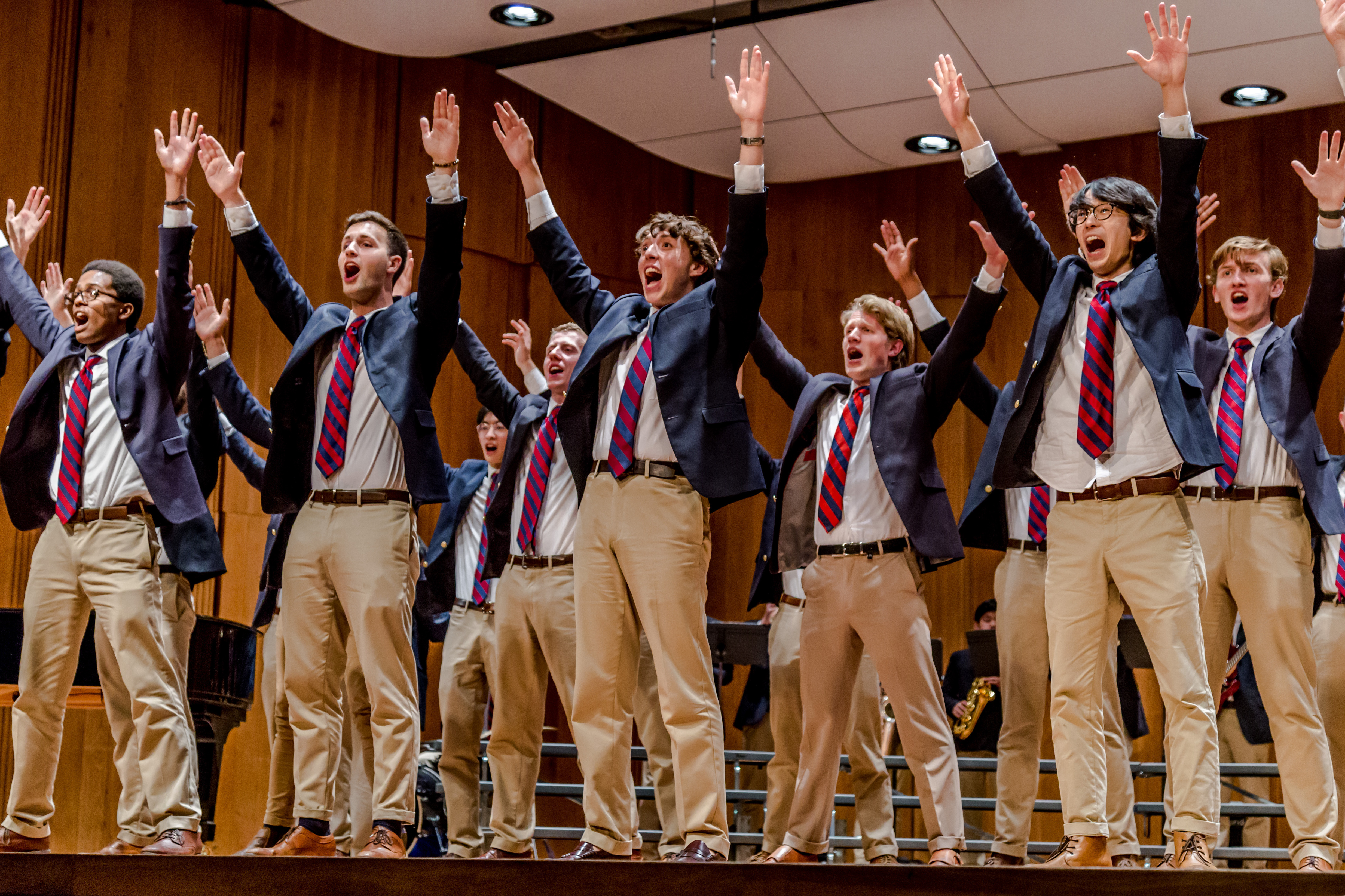 Three lines of students on stage dressed in blue blazers, khaki pants, and red-and-blue-striped ties singing with their arms over their heads. 