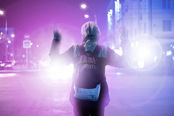 A person with arms raised wearing a backpack at night faces the headlights of a vehicle on a city street.