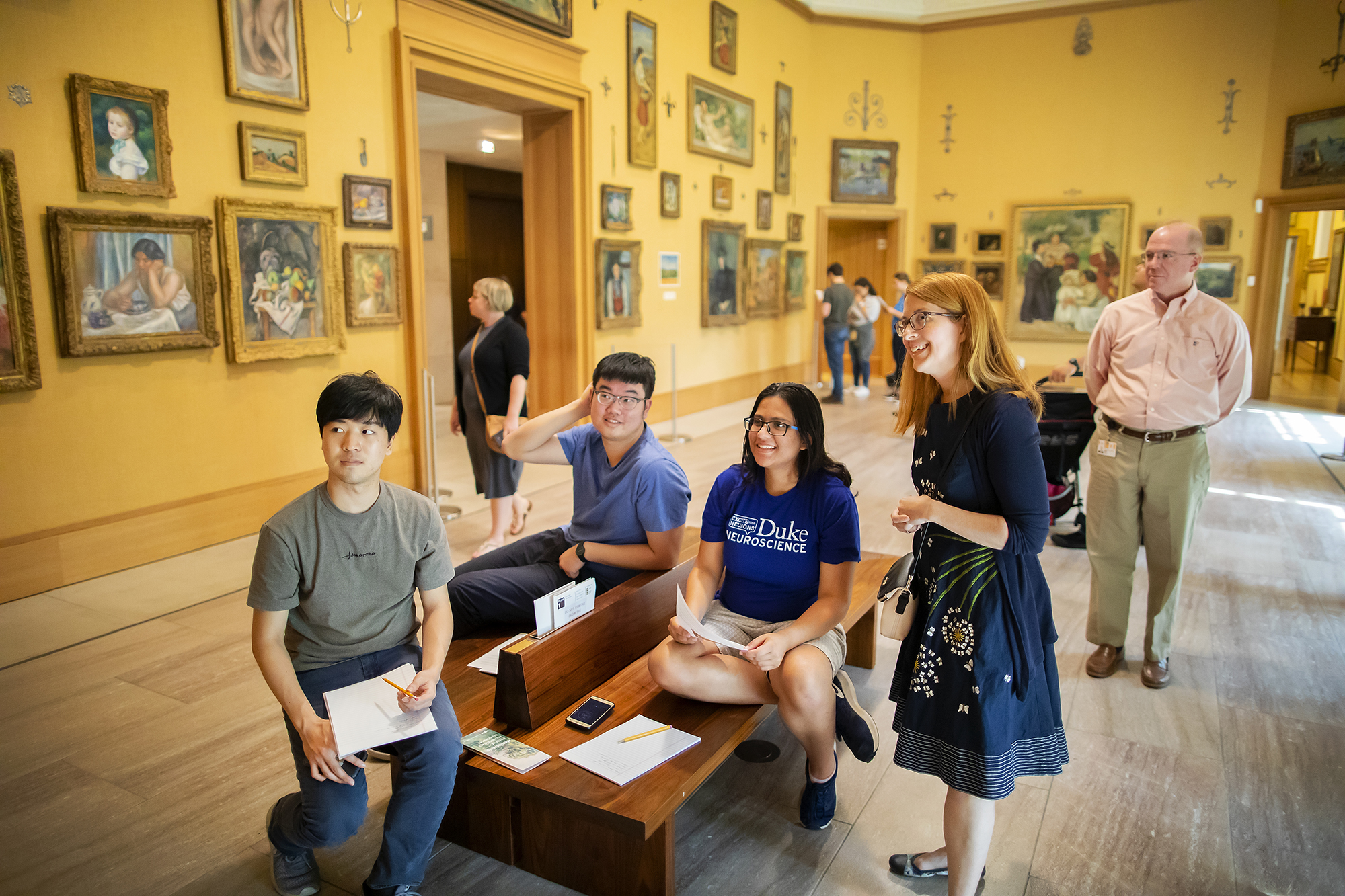 A group of people, some sitting on a bench, some standing, looking at something offscreen, with paintings on yellow walls in the background.