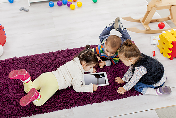 three toddlers on a mat on the floor of a playroom looking at a tablet